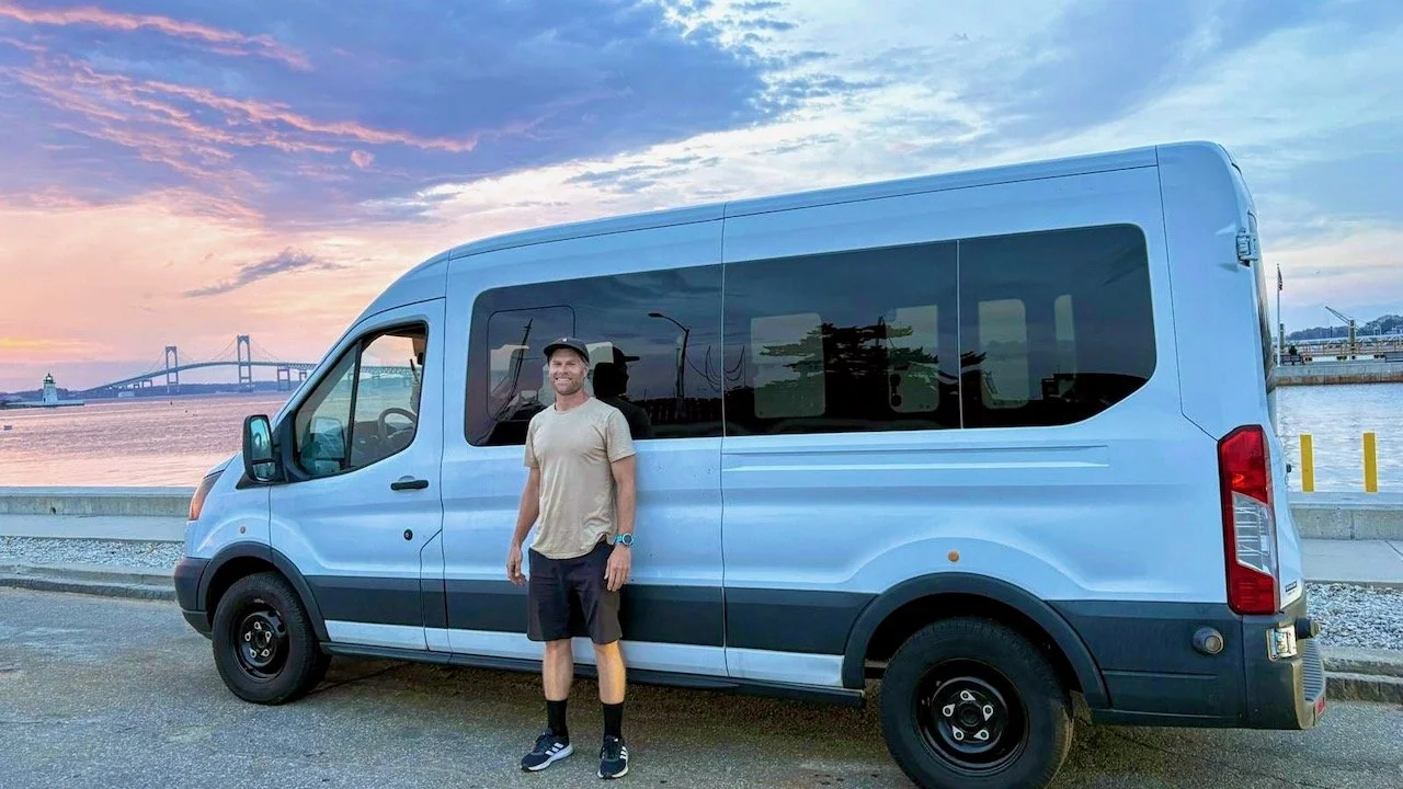 A man stands next to a large white van near a waterfront during sunset, with a bridge and water in the background.