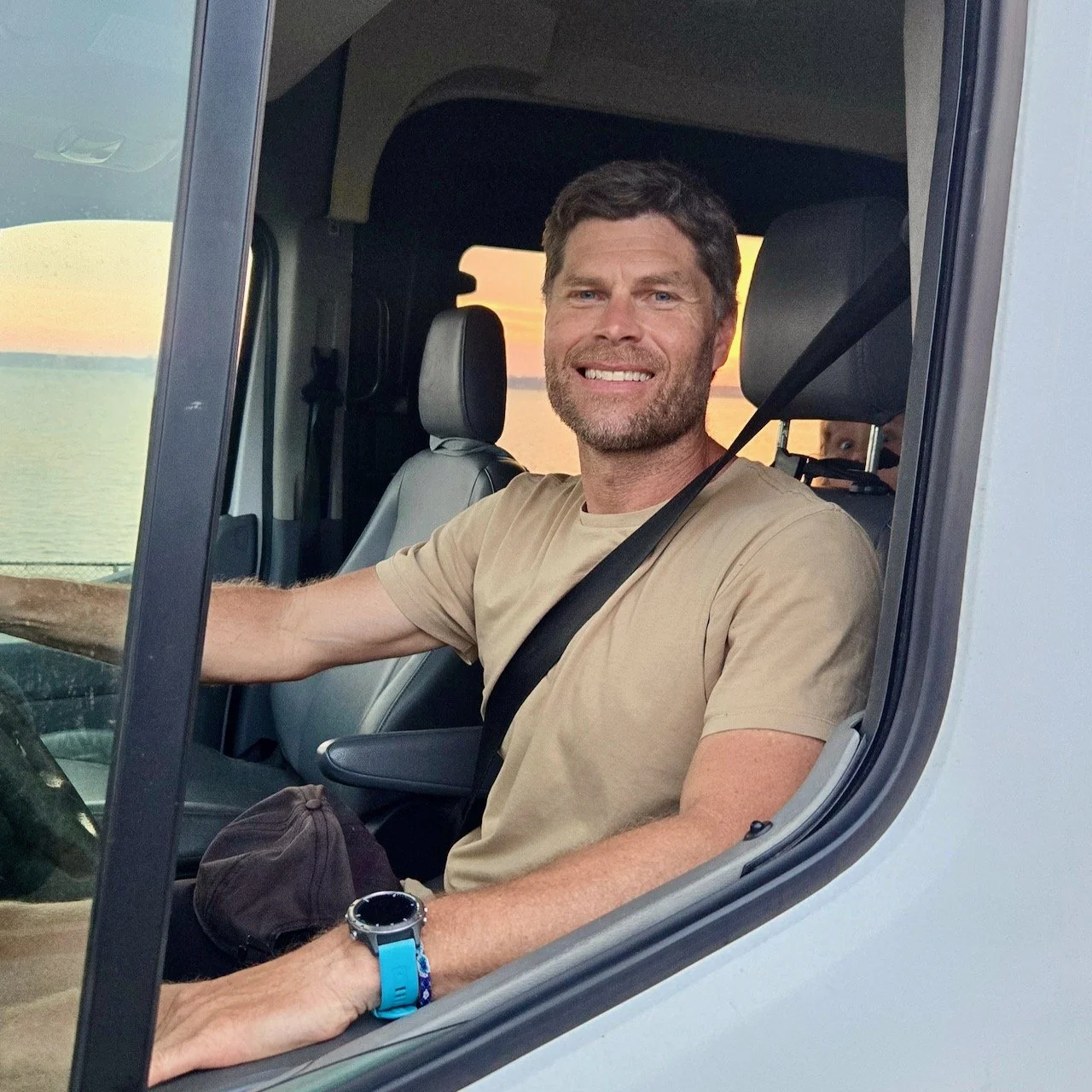 A man with short brown hair and a beard smiling while sitting in the driver's seat of a vehicle, with a sunset over water visible in the background.