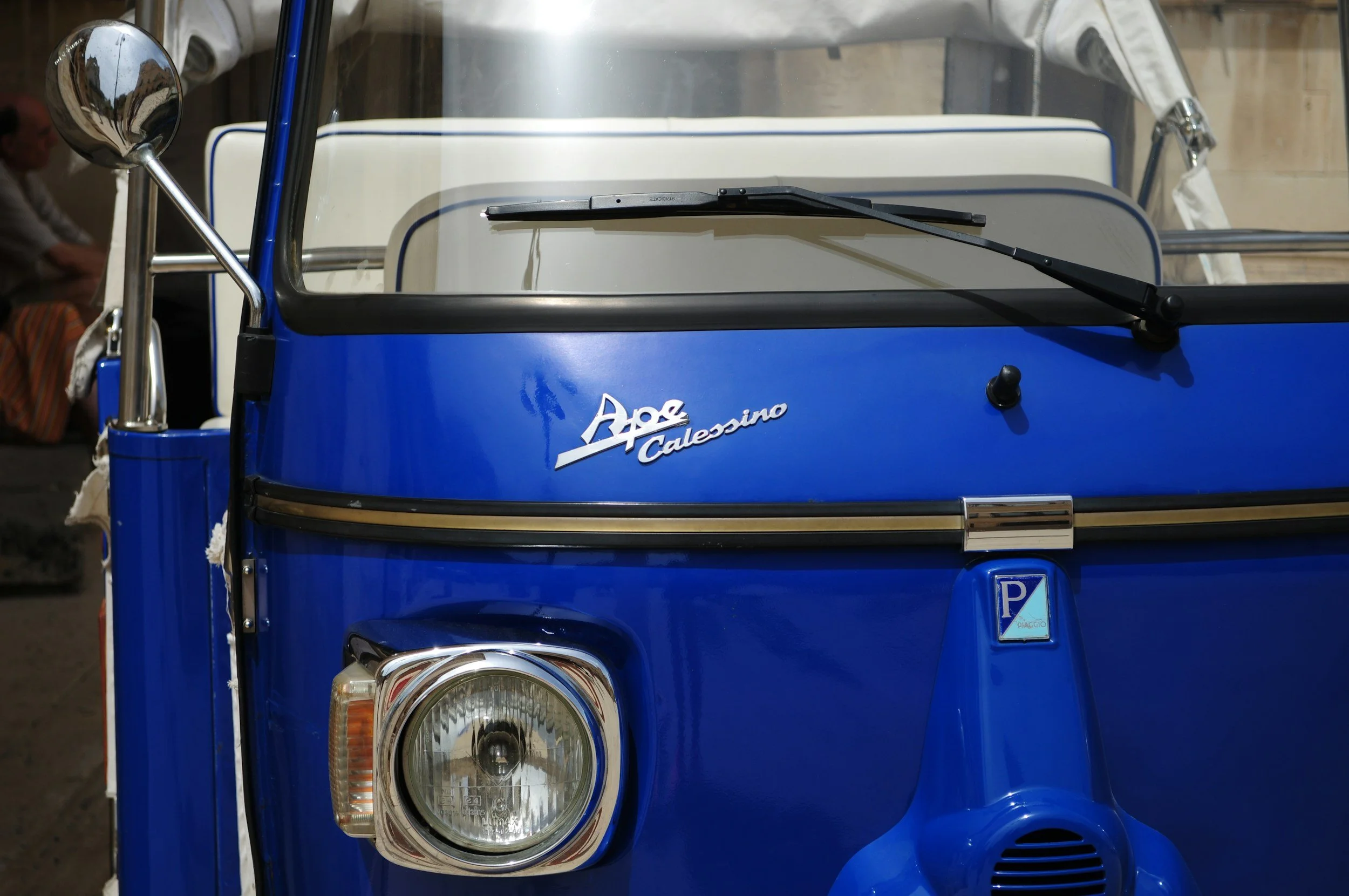 Nahaufnahme eines blauen Piaggio Ape Calessino mit Frontscheinwerfer und Windschutzscheibe.