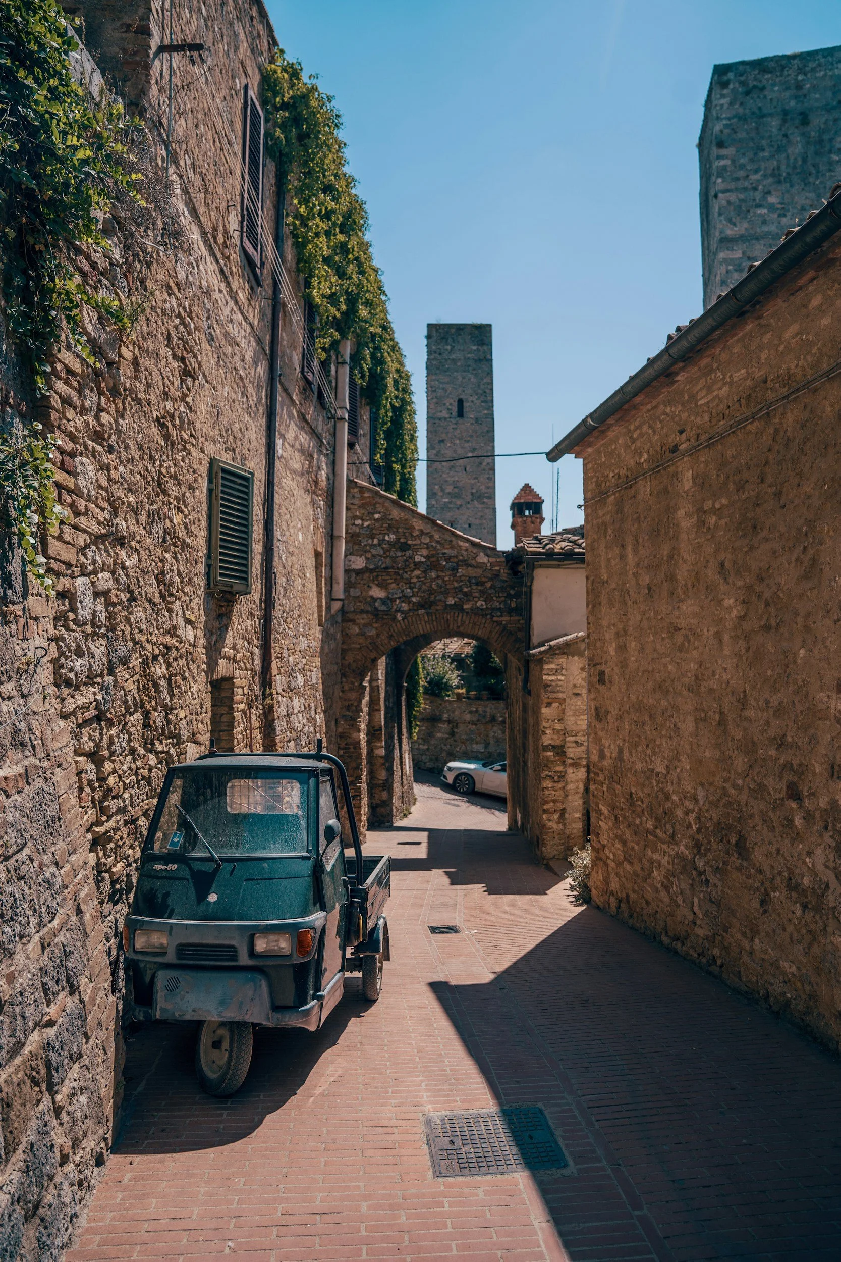Eine enge, historische Gasse mit alten Steinmauern, einem kleinen grünen Traktor und zwei parkenden Autos im Hintergrund, bei sonnigem Wetter.