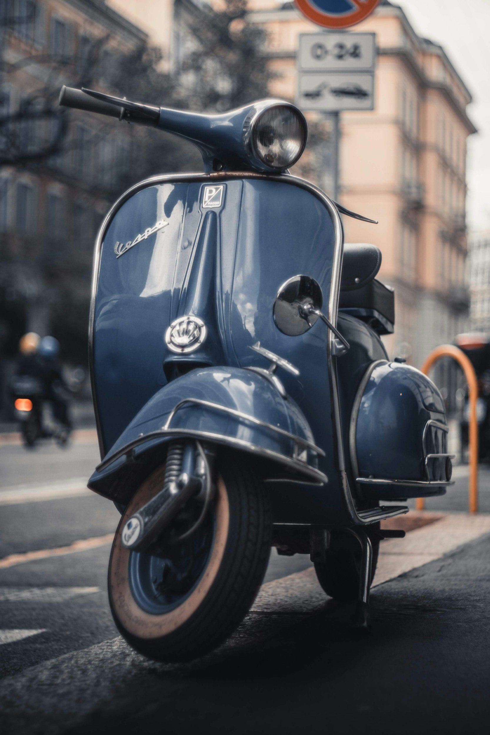 Ein vintage blauer Vespa-Roller parkt auf der Straße, Nahaufnahme, städtische Umgebung, im Hintergrund Gebäude und andere Roller.