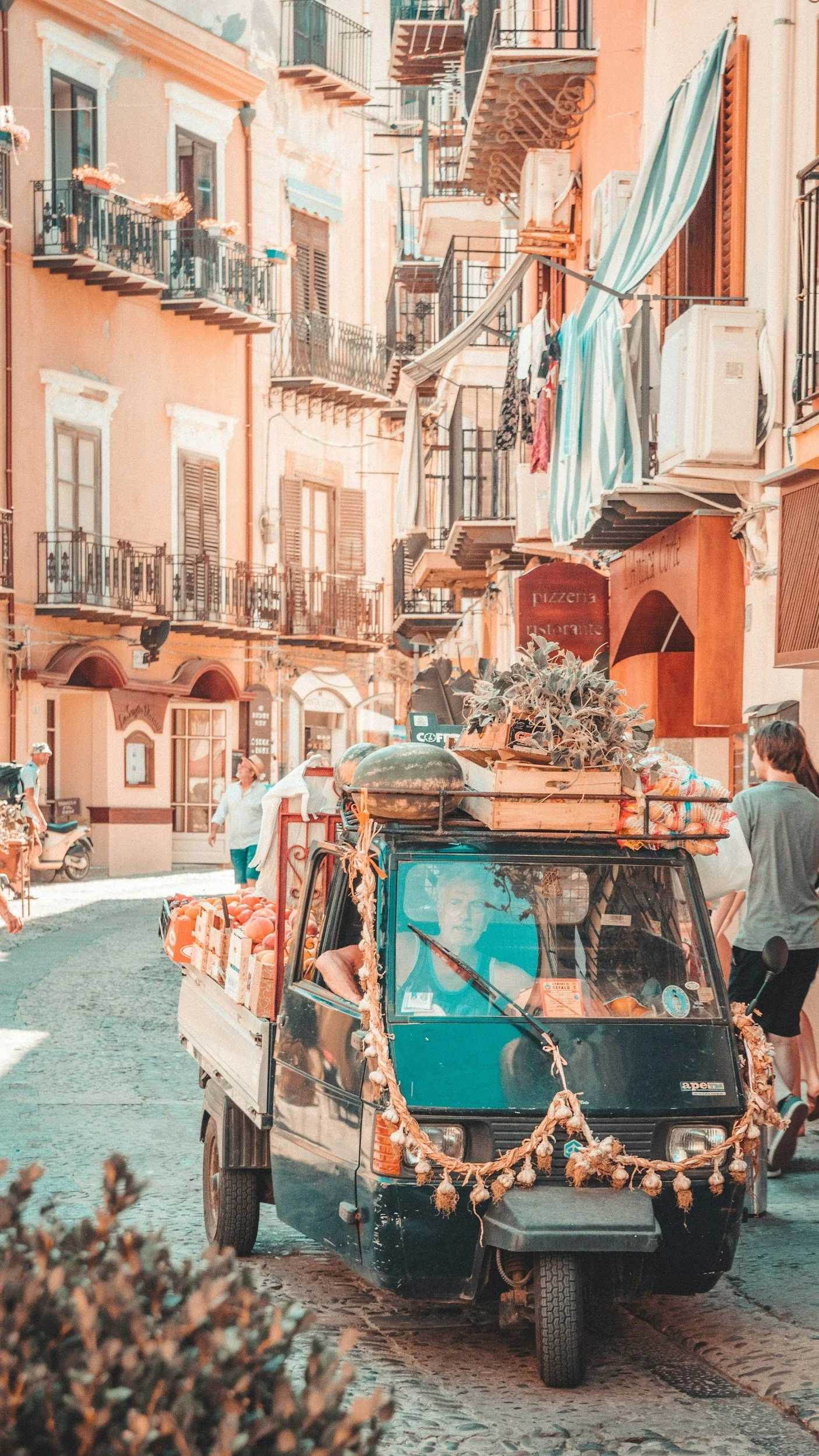 Ein kleines grünes Auto, das auf einer schmalen europäischen Straße geparkt ist, mit einem Dach voller Pflanzen und Obst, umgeben von bunt bemalten Gebäuden mit Balkonen und Wäsche, die im Wind weht, und einigen Menschen, die vorbeigehen.