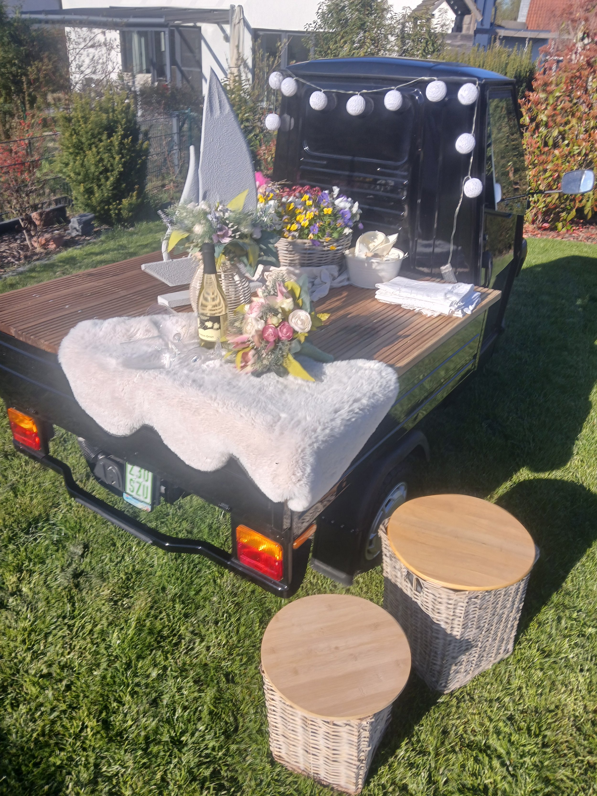 Ein kleines schwarzes Dreirad mit Holzplatte, auf der Pflanzen, Blumen, eine Weinflasche, Geschirr und eine Kerze stehen, dekoriert mit weißen Papierlaternen, im grünen Garten.