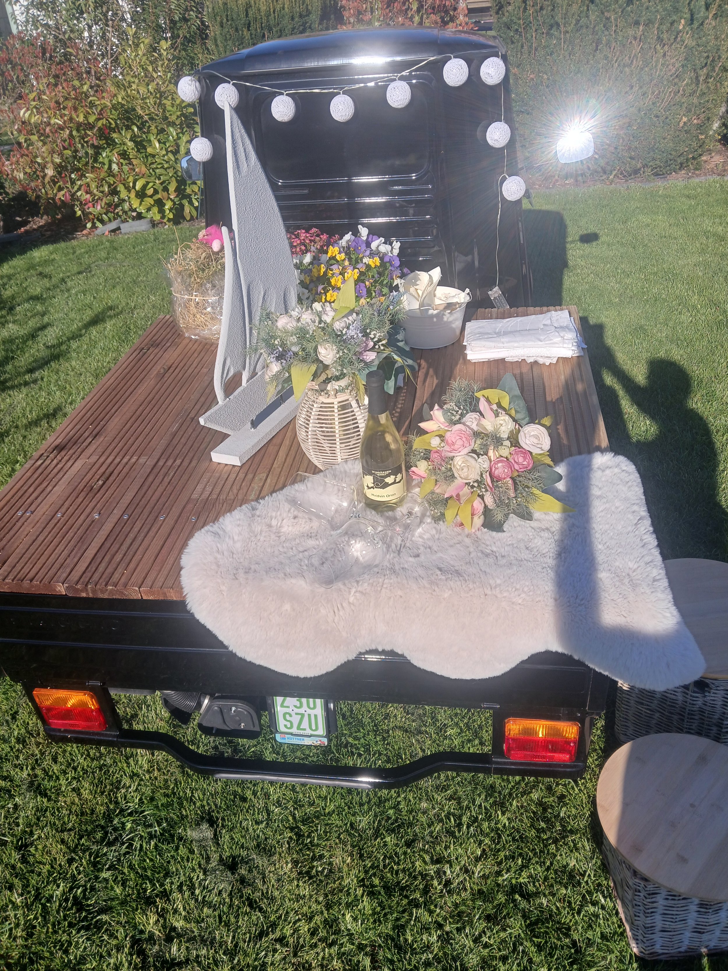 Auf einem kleinen Anhänger sind Blumen, eine Flasche Weißwein, Dekogegenstände, und Geschenke arrangiert, dekoriert mit weißen Lichterketten, im Hintergrund ist eine grüne Rasenfläche und ein Gebüsch zu sehen. Die Sonne scheint stark, was eine Reflex
