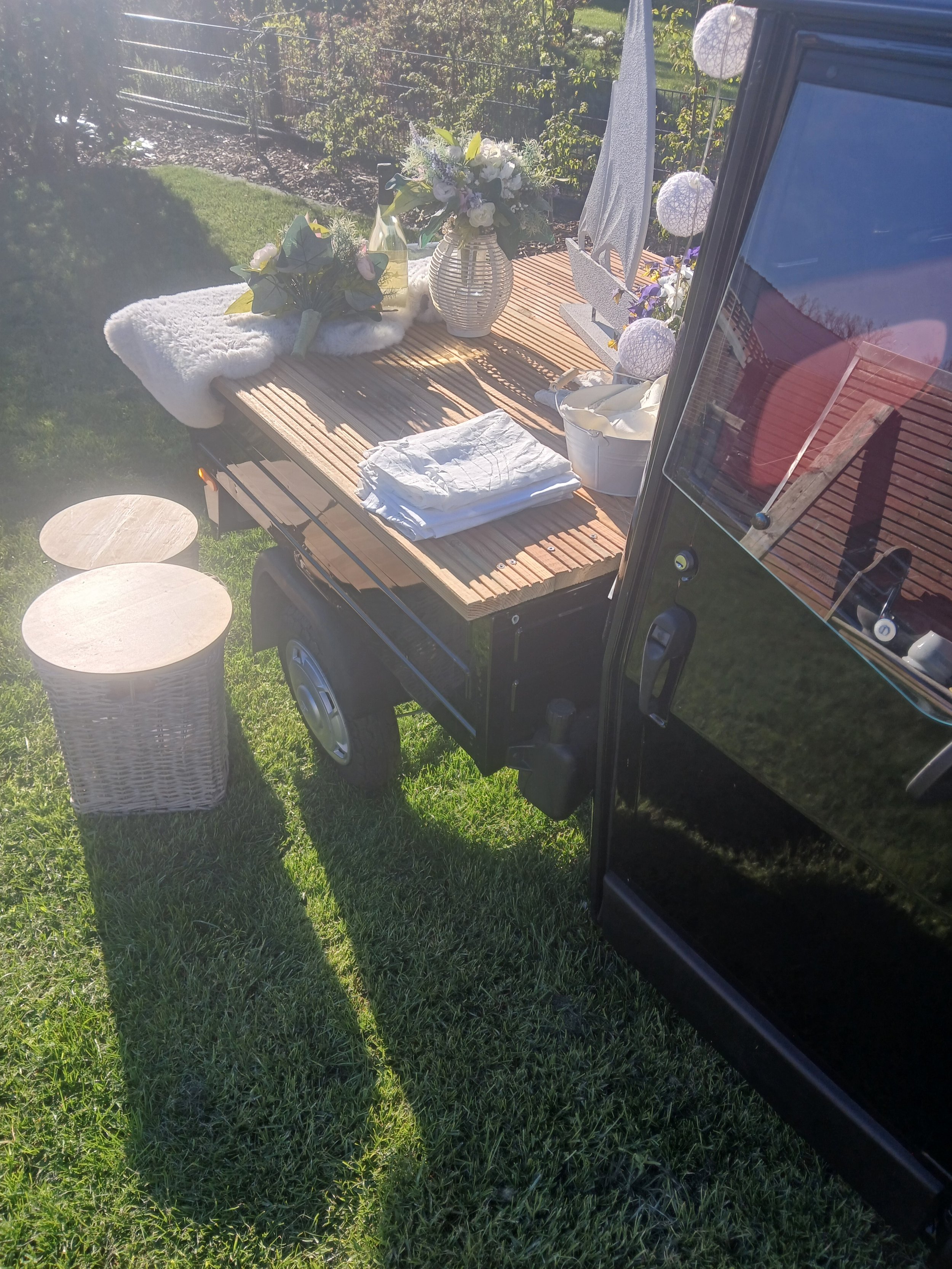 Ein Garten während des Tages mit einem Wagen, auf dem eine Tischdekoration mit Blumen und Lampions steht. Es gibt zwei kleine runde Tische auf dem Rasen, ein gefaltetes Handtuch liegt auf der Tischplatte des Wagens, und eine kleine weiße Kiste mit St