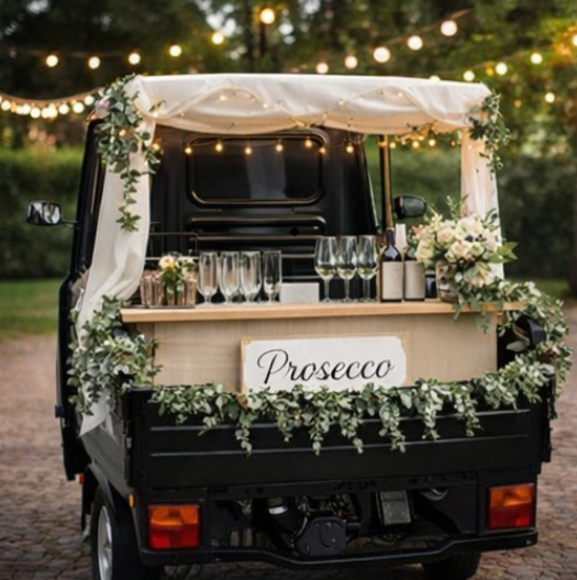 Ein food truck, der mit Champagnerglas und Blumen dekoriert ist, mit einem "Prosecco"-Schild, umgeben von Lichterketten im Freien, wahrscheinlich bei einer Feier oder Veranstaltung.