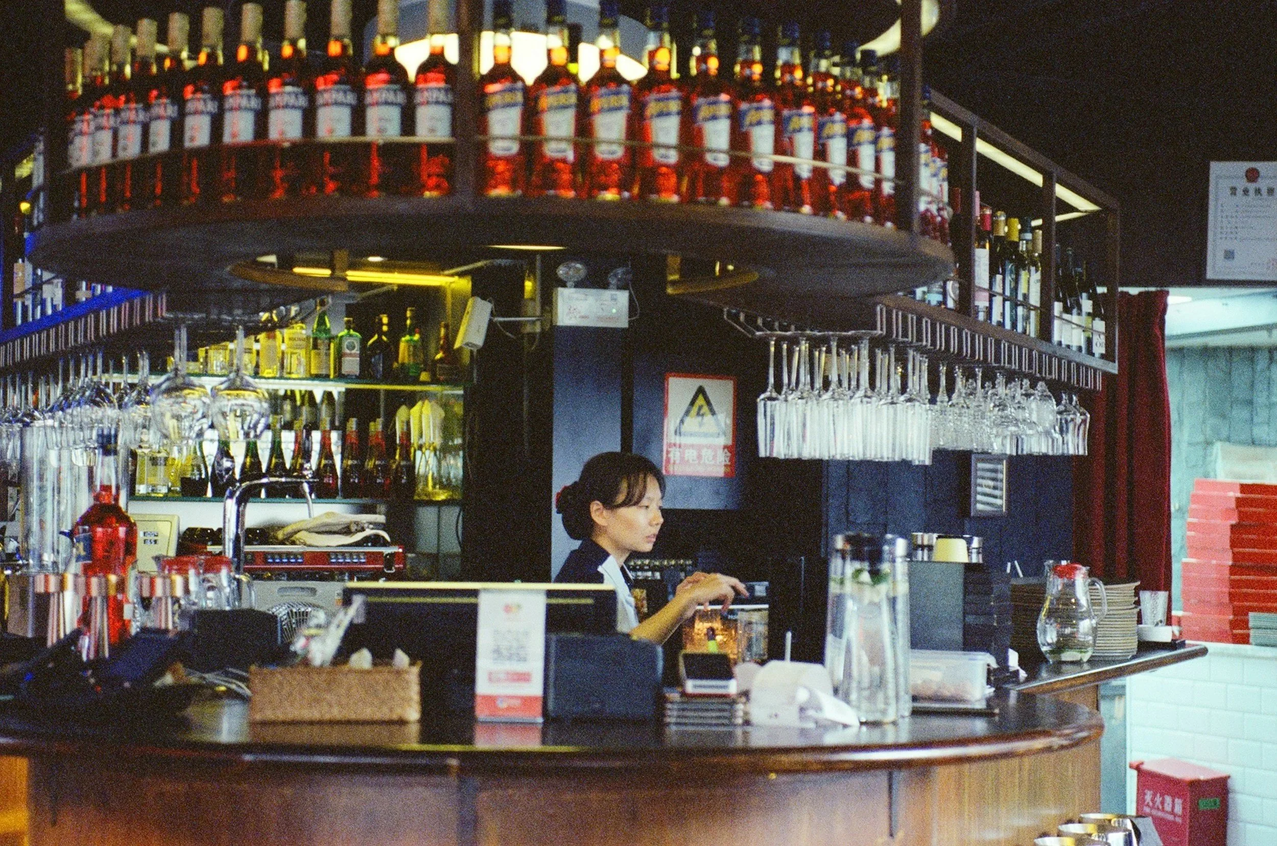 Bar mit einer Barkeeperin hinter dem Tresen, umgeben von Flaschen und Gläsern, mit Rotweinflaschen im oberen Regal und verschiedenen Spirituosen im Hintergrund, in einem dimmbeleuchteten Lokal.