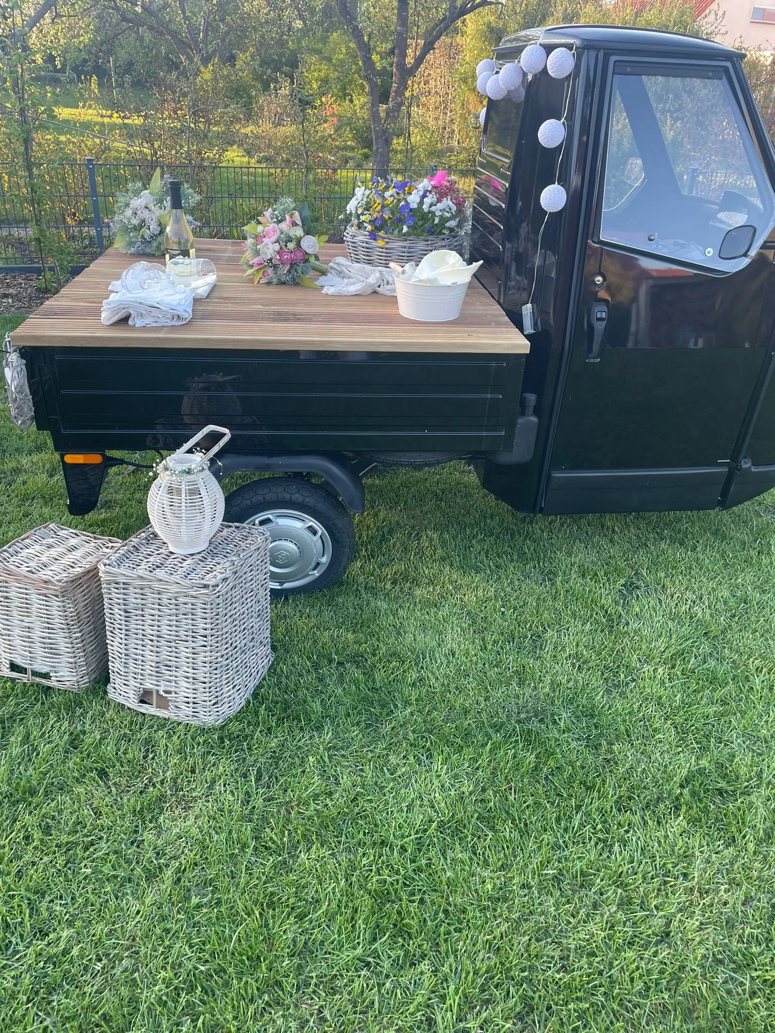 Ein schwarzer rollbarer Wagen mit einer Holzplanke auf der Ladefläche, dekoriert mit Blumen, einer Flasche Wein und einem Gläser. Vor dem Wagen befindet sich ein Korb, in dem eine weiße Laterne steht, auf dem Rattanhocker. Im Hintergrund ein Garten m