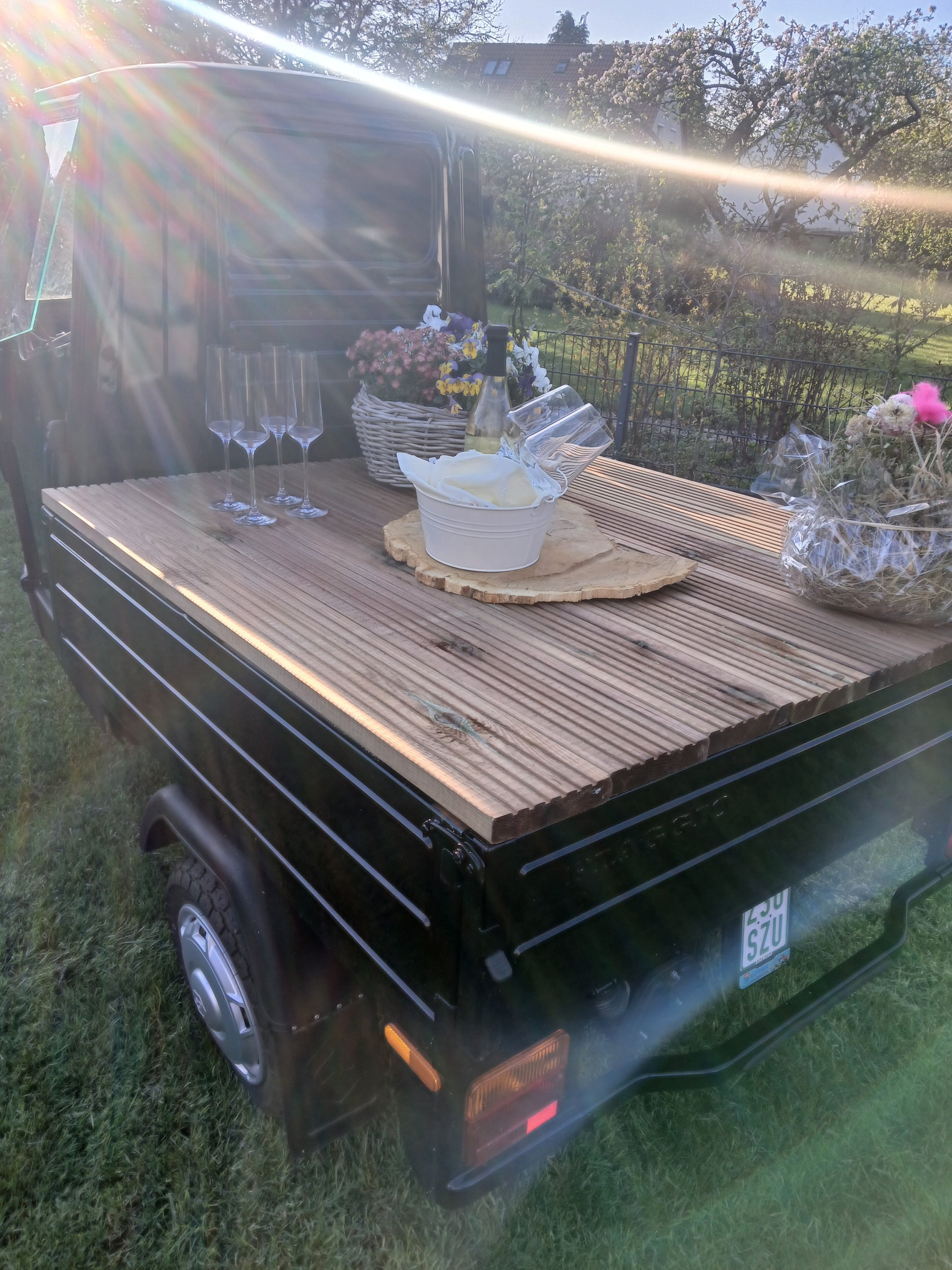 Ein mobiler Grillwagen mit einer Holzarbeitsfläche, auf der Sektgläser, eine Flasche Wein, Blumen und andere Gegenstände stehen, im Freien bei Sonnenlicht.