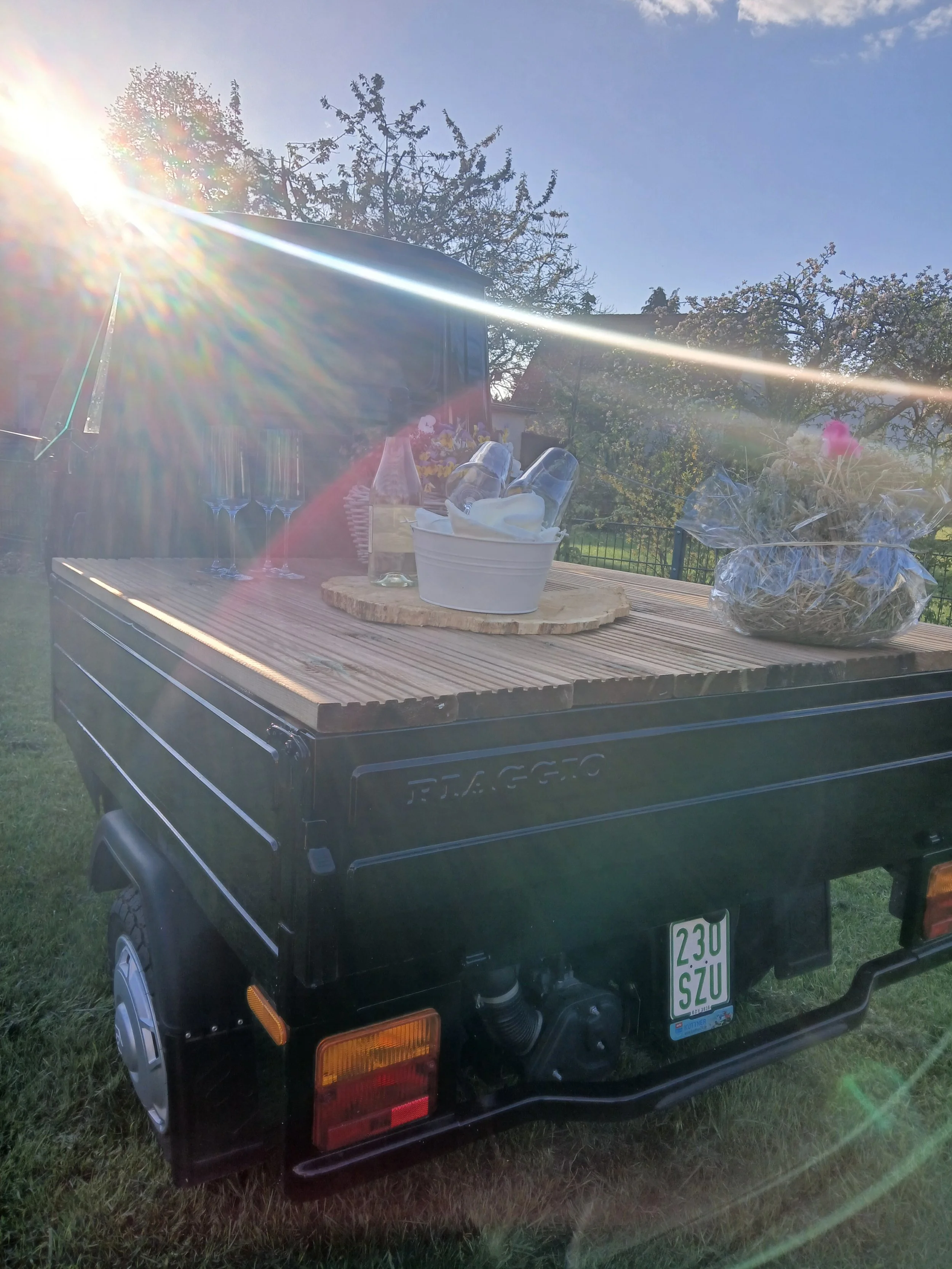 Ein schwarzer Anhänger mit Holztisch, Gläsern und Blumen ist im Garten bei Sonnenlicht.