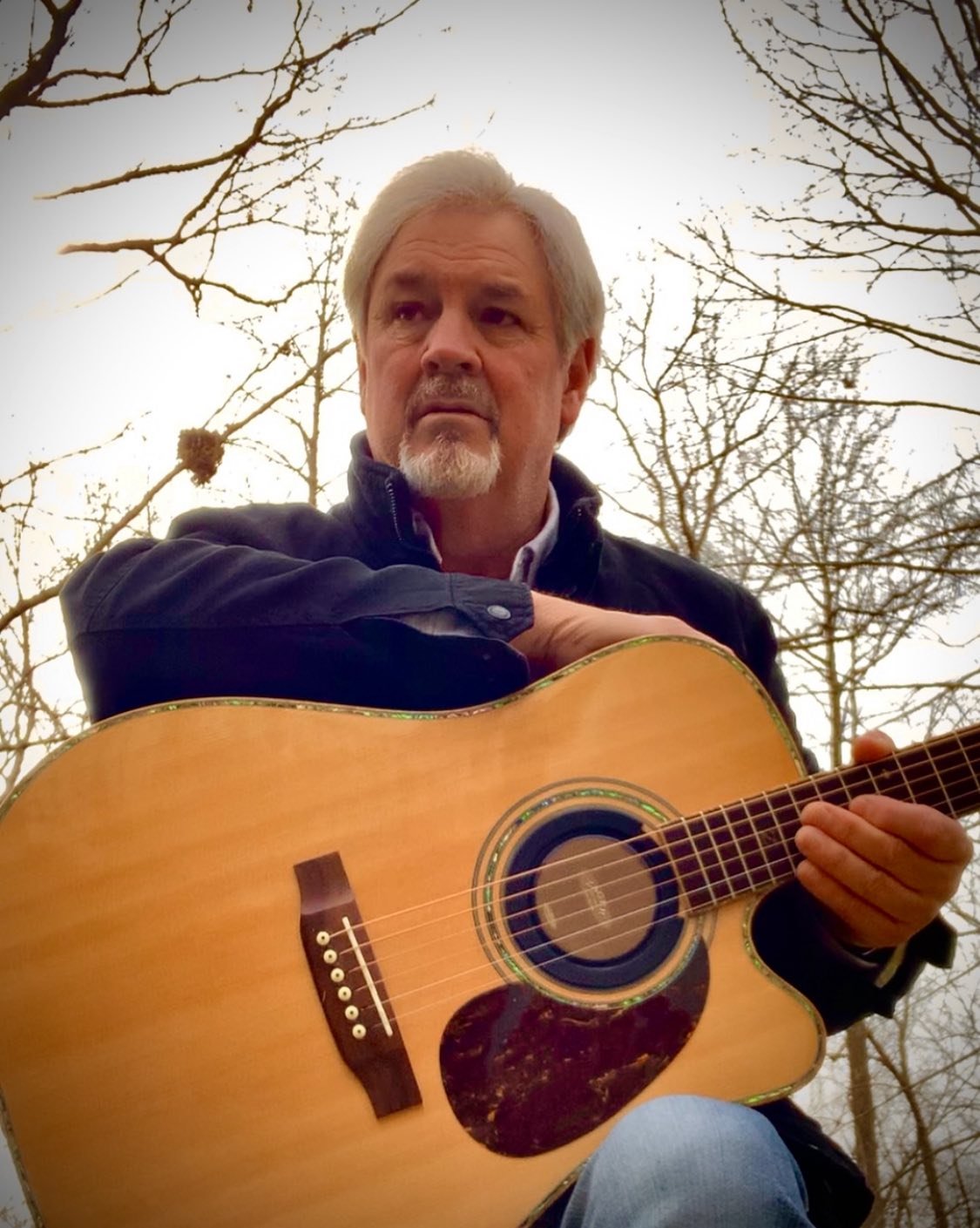 A man with gray hair and a goatee playing an acoustic guitar outdoors, with bare tree branches and a bright sky in the background.