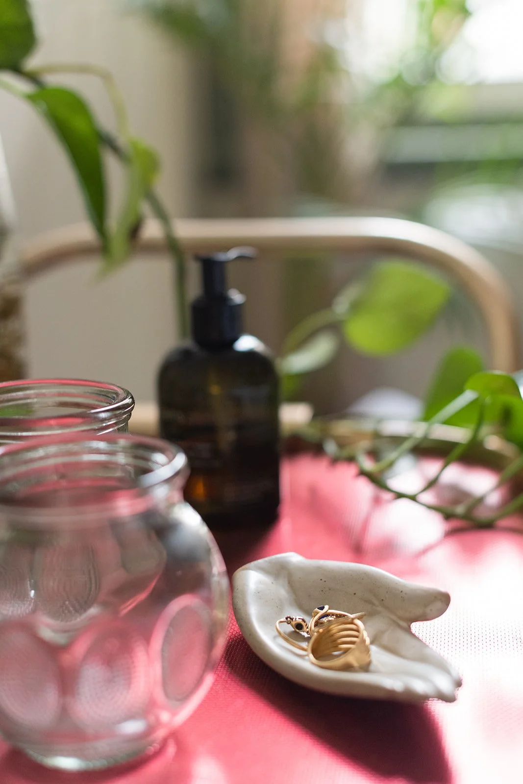 Close-up of a pink table in a massage therapy room with jewelry on a ceramic dish, cupping therapy tools, green leaves, and a black massage oil dispenser, with a blurred background of a window and green plants.