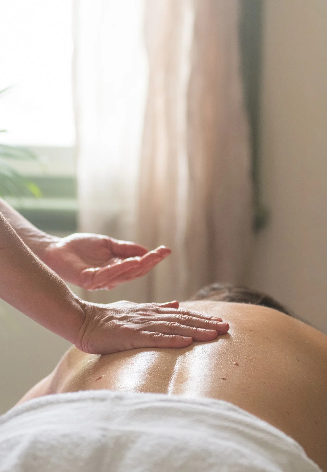 A person receiving a massage on their upper back pain in a bright, softly lit room with curtains and a window.
