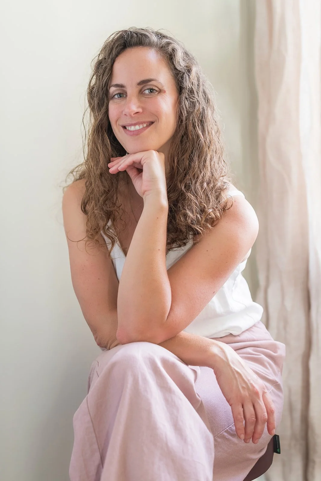 A woman with curly brown hair, smiling, with her hand resting on her chin, sitting indoors near a light-colored curtain.