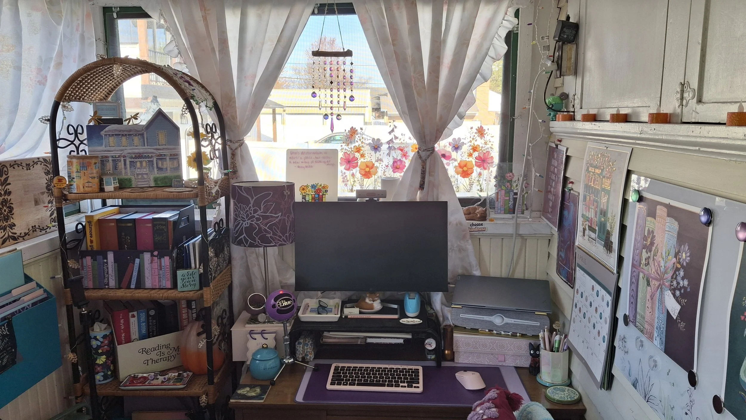 A cozy workspace near a window with floral curtains, decorated with floral window decals, picture frames, books, a computer monitor, and various decorative items.