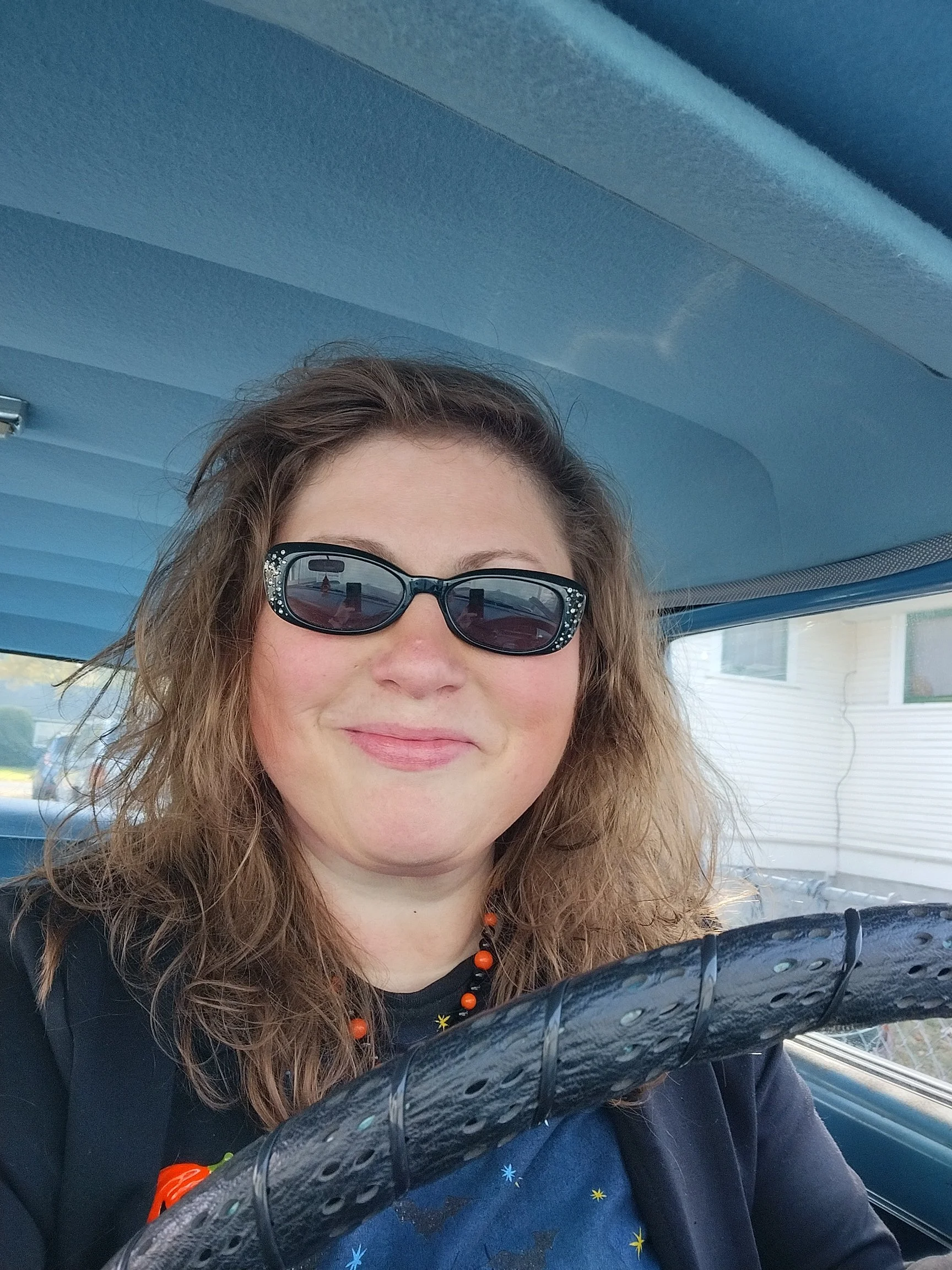 A woman with curly brown hair wearing glasses with decorative rhinestones, a black shirt with star patterns, and a necklace with orange beads is sitting in a vehicle. The photo captures part of her face and upper body with a view of the vehicle's interior and the house visible outside through the window.