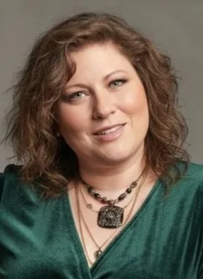 A woman with curly brown hair wearing a green top and layered necklaces, smiling softly.