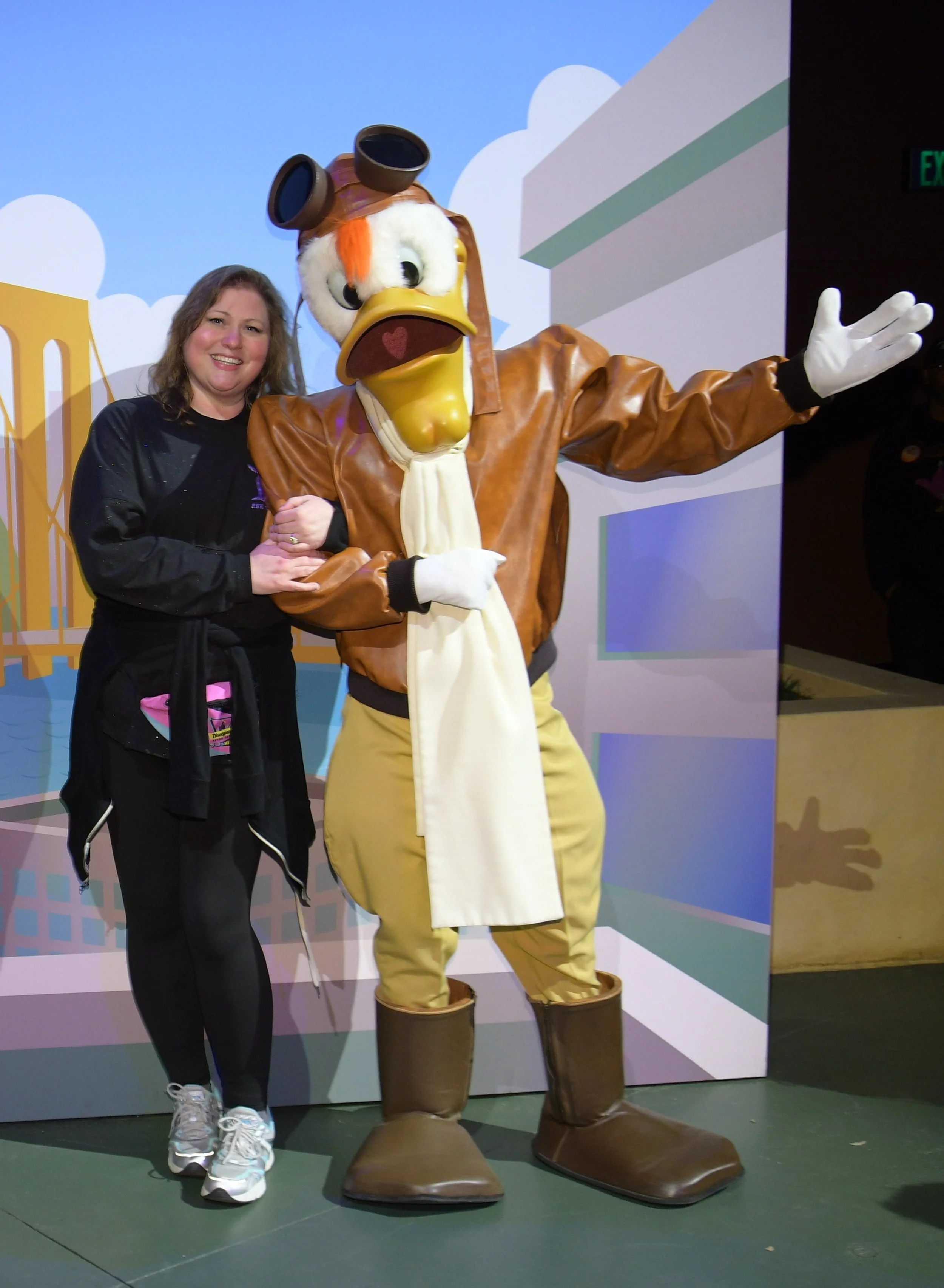 A woman standing next to a life-sized mascot of Launchpad McQuack wearing a brown leather jacket, yellow pants, and large boots, with a colorful background depicting a cartoon cityscape.
