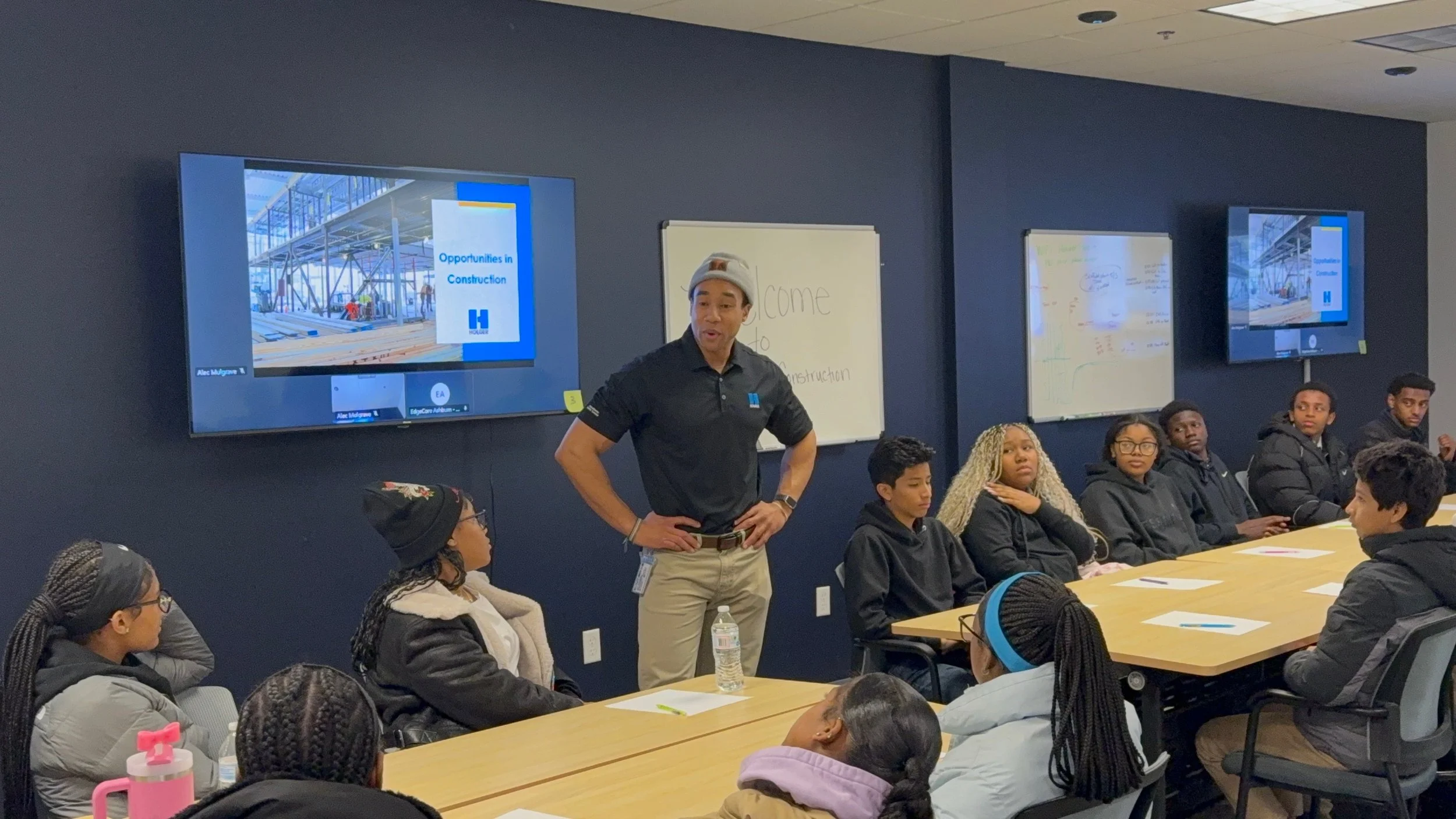 A man stands at the front of a classroom filled with students, presenting about construction opportunities. A large screen displays an image of a construction site with workers and the text 'Opportunities in Construction'. Students are seated around a long table, attentively listening.