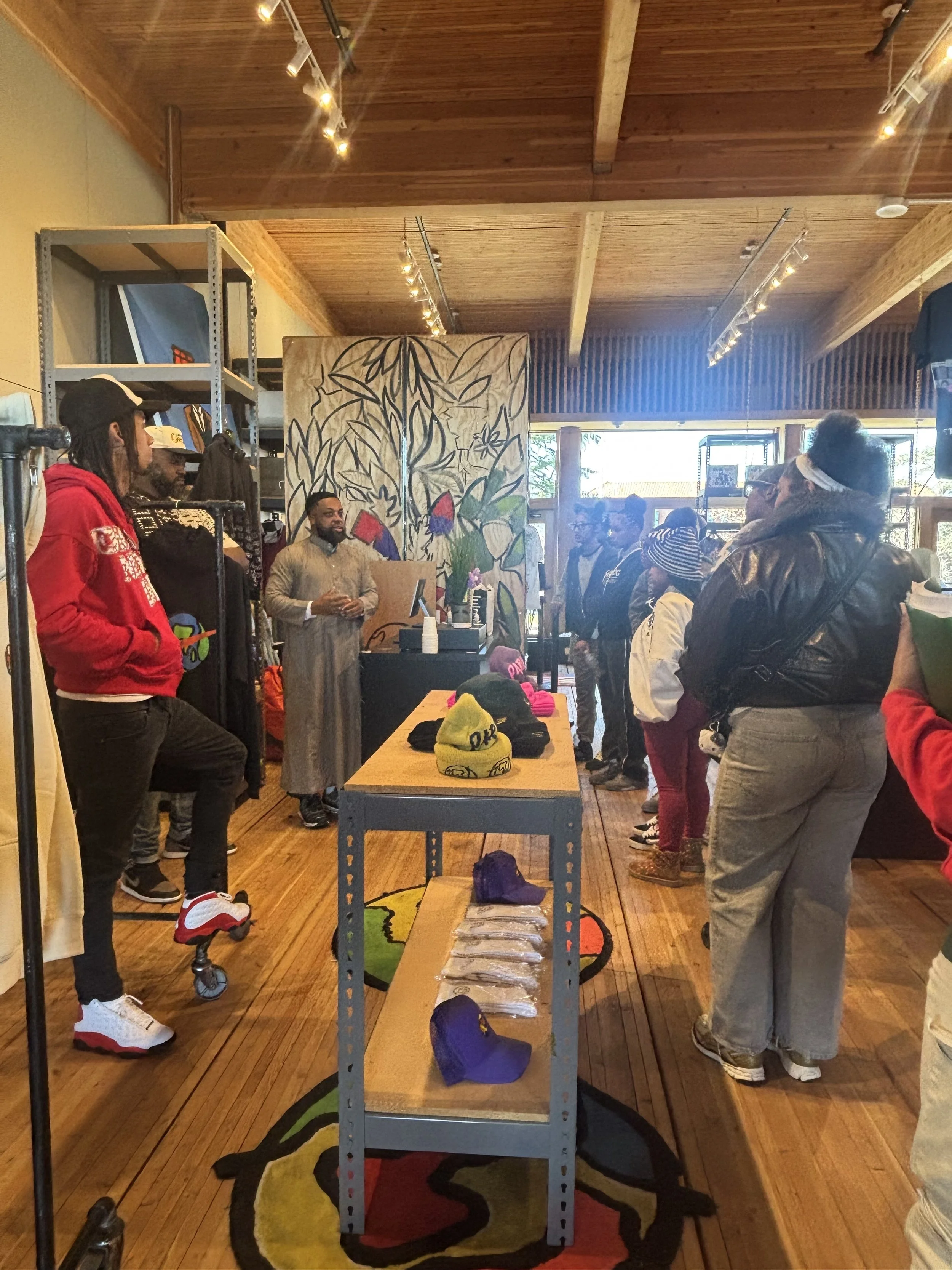 Group of people gathered inside a store or gallery, some standing and some sitting, with merchandise and artwork displayed, and a large colorful mural on the wall behind them.