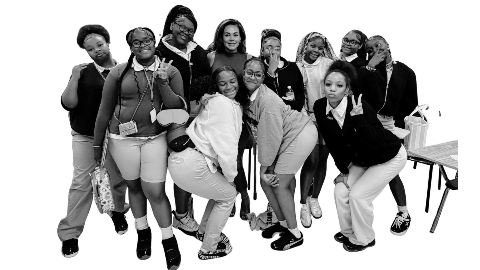 Black and white photo of a diverse group of young women posing together indoors, smiling and making peace signs, with some hugging and leaning on each other.