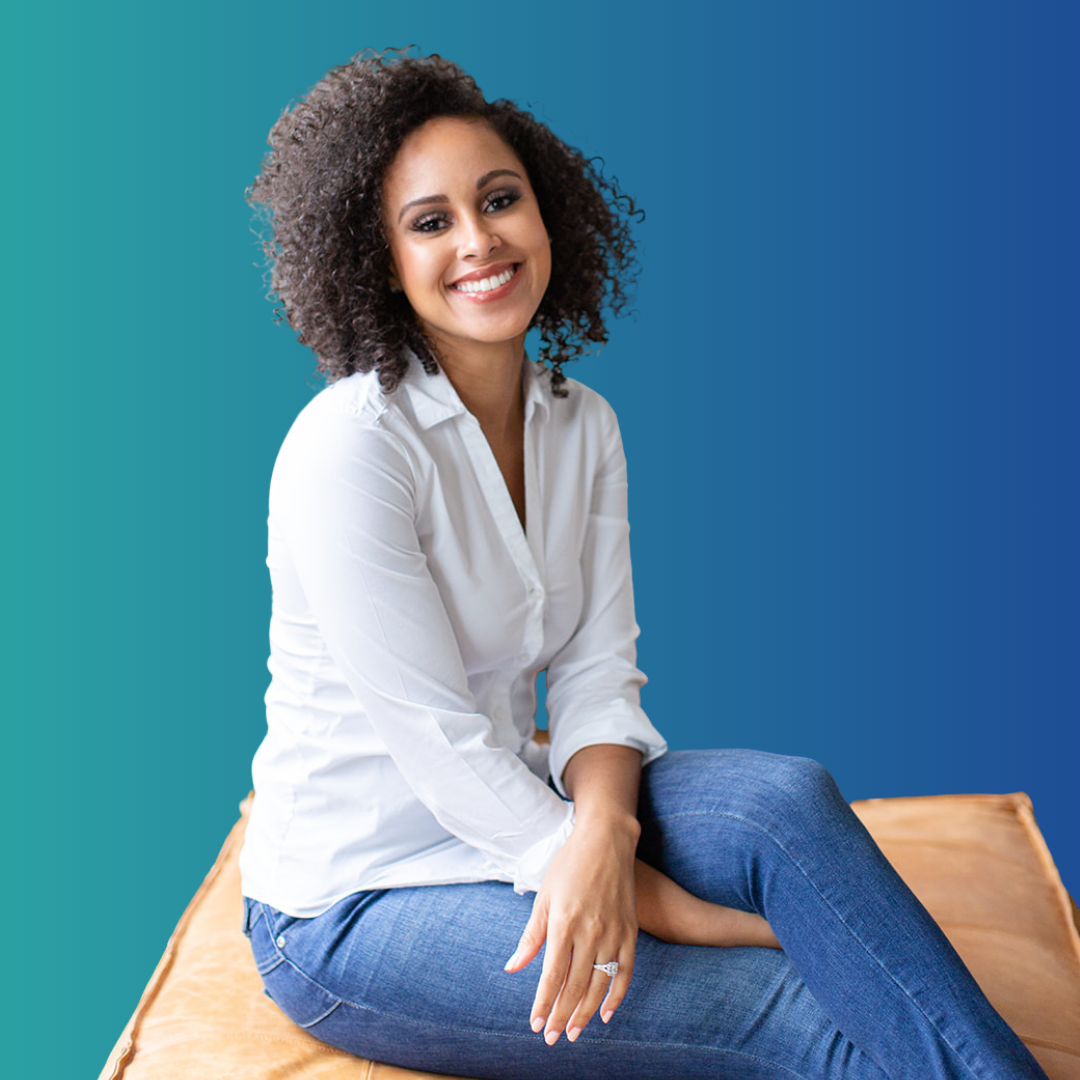 Smiling woman with curly hair, wearing a white shirt and jeans, sitting on a wooden surface with a gradient blue background.