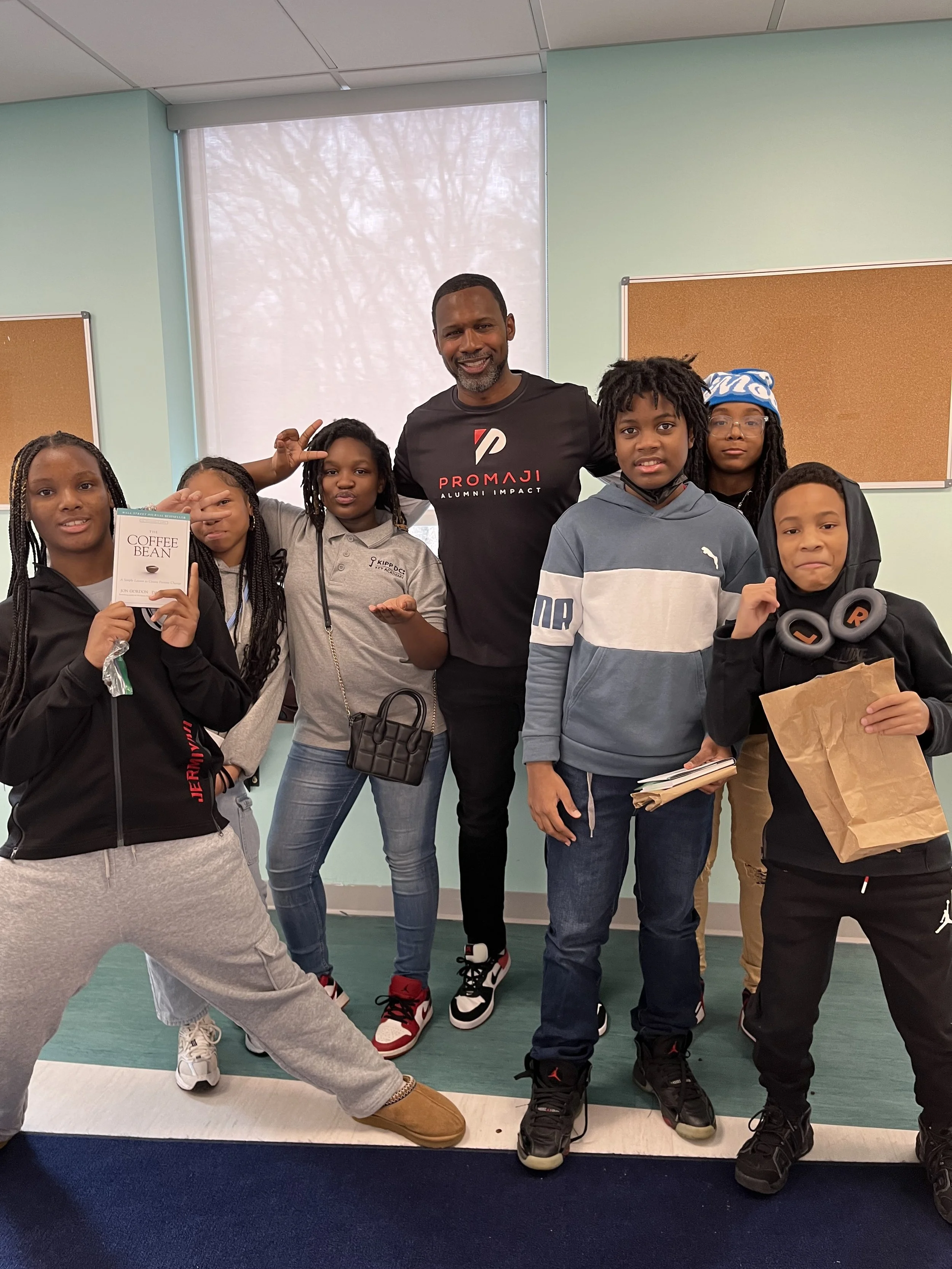 A group of six children and one adult posing together indoors, with a large window behind them. The children are making various hand gestures and holding items such as a book, paper bag, headphones, and a small backpack. The adult is standing in the middle, smiling and wearing a black t-shirt with red and white text. The room has light green and beige walls, and the children are dressed casually.