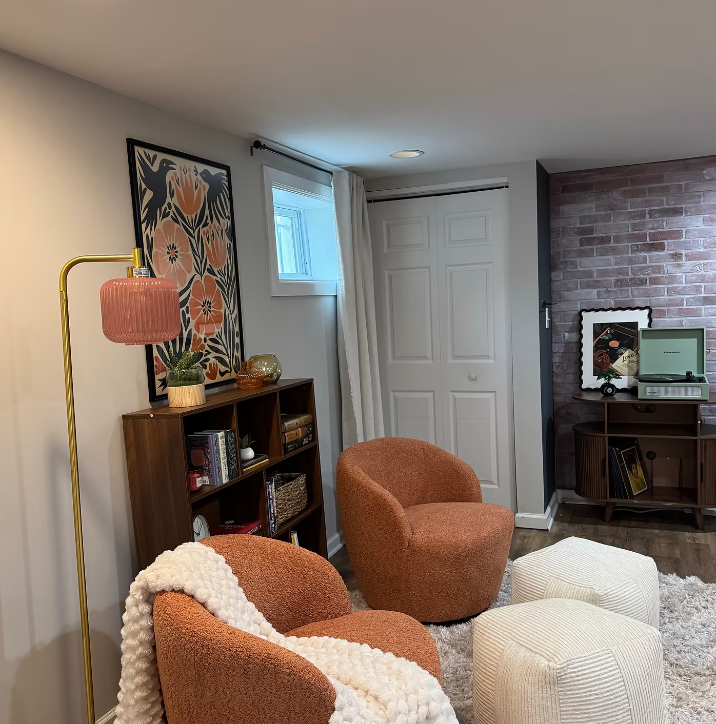 A cozy living room corner with orange mid-century modern chairs, a white textured pouf, a shaggy area rug, a wooden bookshelf with decorative items and books, a framed floral art piece, a pink table lamp with a gold stand, a small window with white curtains, and a brick accent wall featuring a framed picture and a vintage record player.