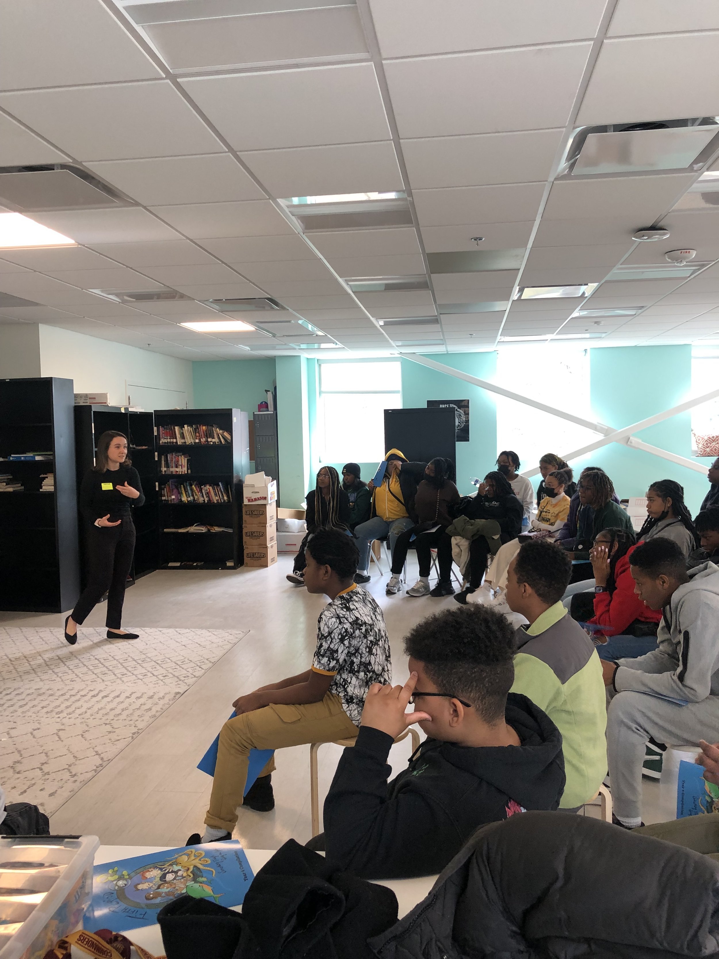 A woman in black clothes is speaking to a group of students seated and standing, in a room with bookshelves and boxes. Students are paying attention, some taking notes, with a bright window behind them.