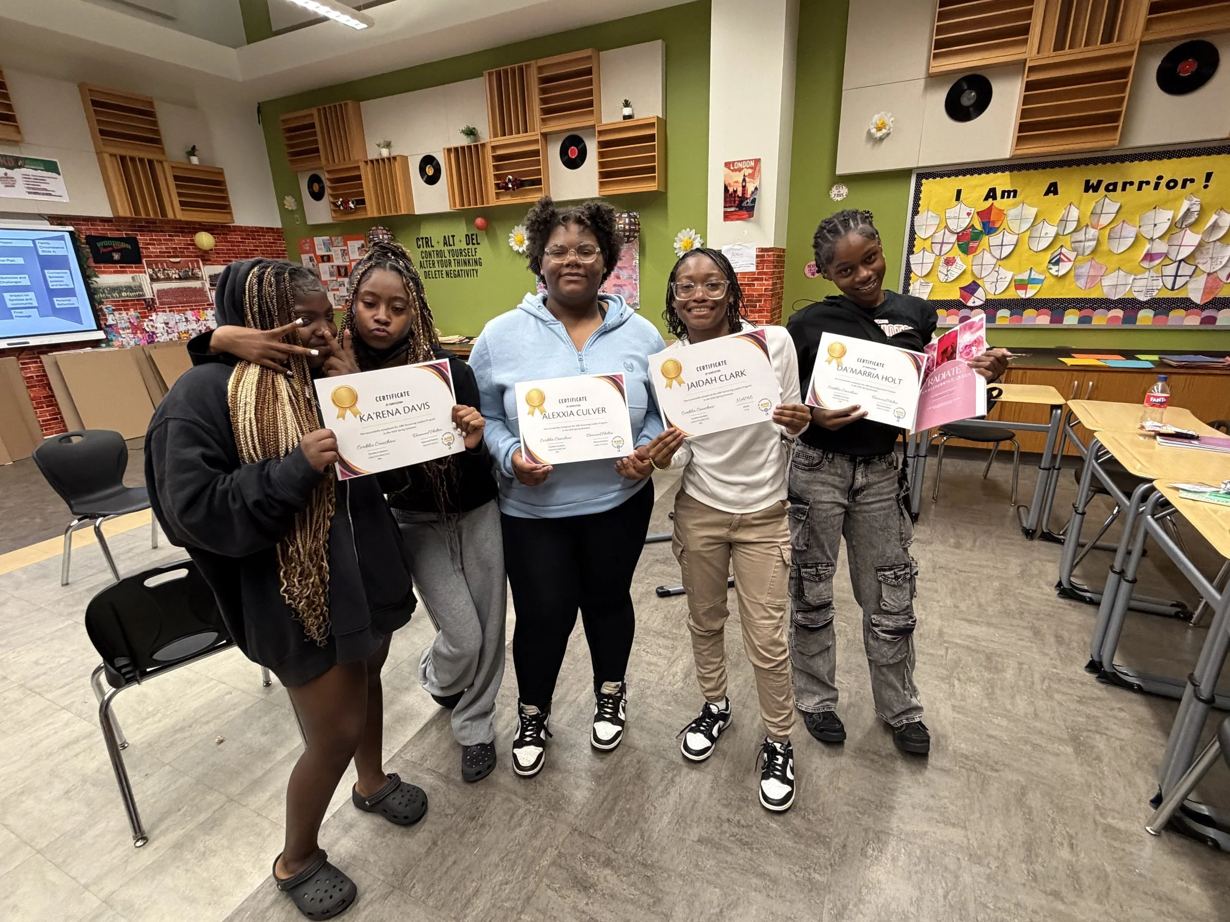 Five young girls standing in a classroom, holding certificates of achievement, smiling, and posing for a photo.
