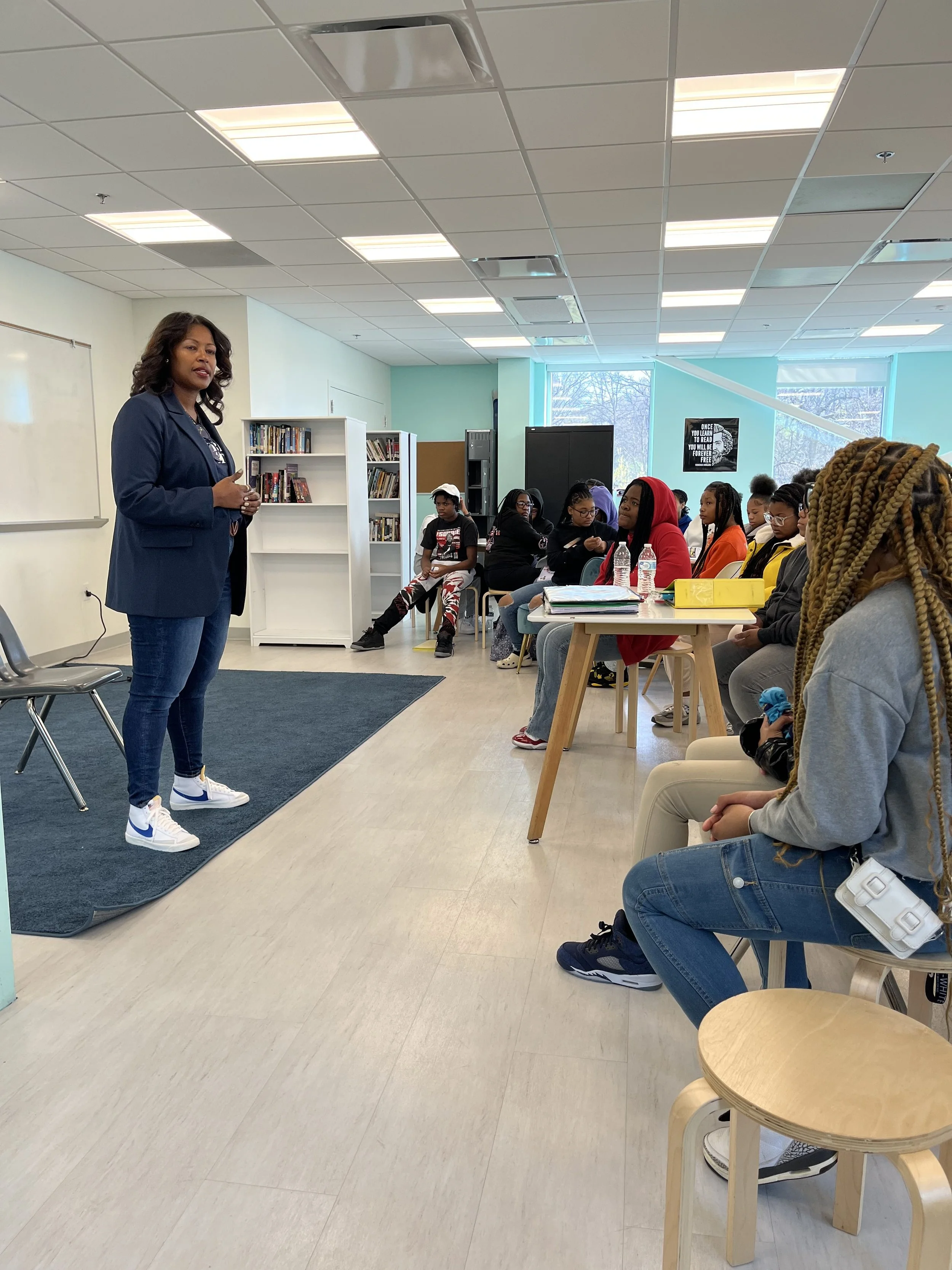 A woman in a navy blazer and jeans standing in front of a classroom speaking to a group of students seated at desks. The students are listening attentively, some taking notes, in a room with bookshelves and large windows.