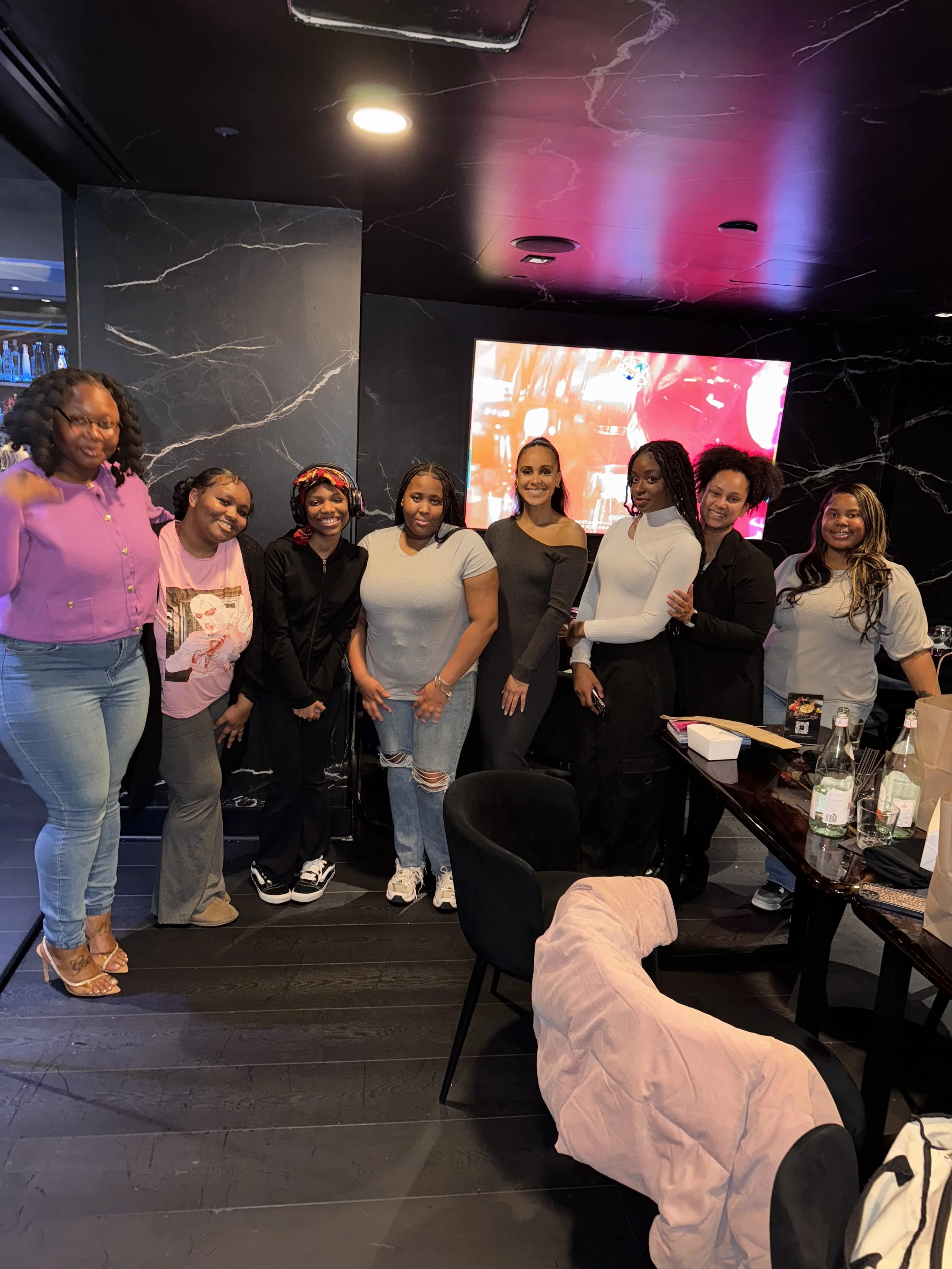 A group of nine women posing together inside a dark, modern room with a black marble wall and a large TV screen in the background. Some women are smiling, and they are dressed casually. There are bottles and papers on the table in front of them, and a pink jacket is draped over a chair in the foreground.