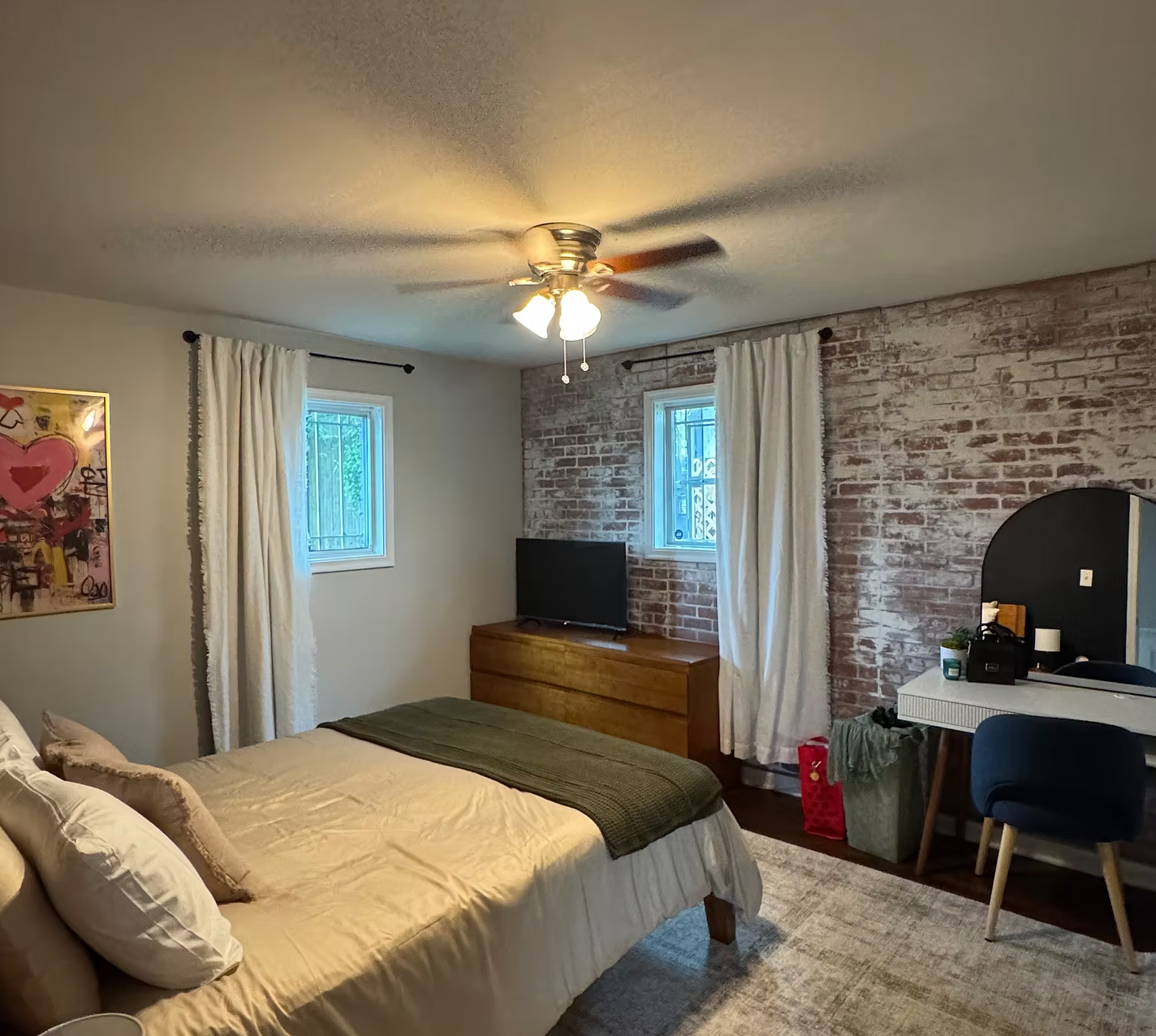 A bedroom with a bed, two windows with white curtains, a ceiling fan with lights, a TV on a wooden dresser, a large art piece on the wall, a white desk with a mirror and black chair, and a brick accent wall.