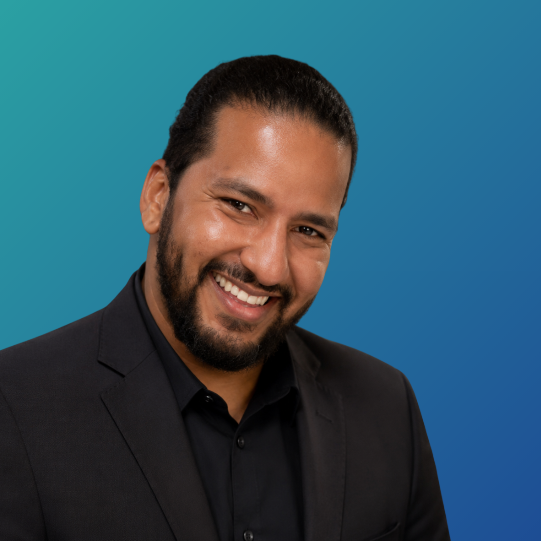 Smiling man with dark hair and beard wearing a black suit against a blue gradient background.