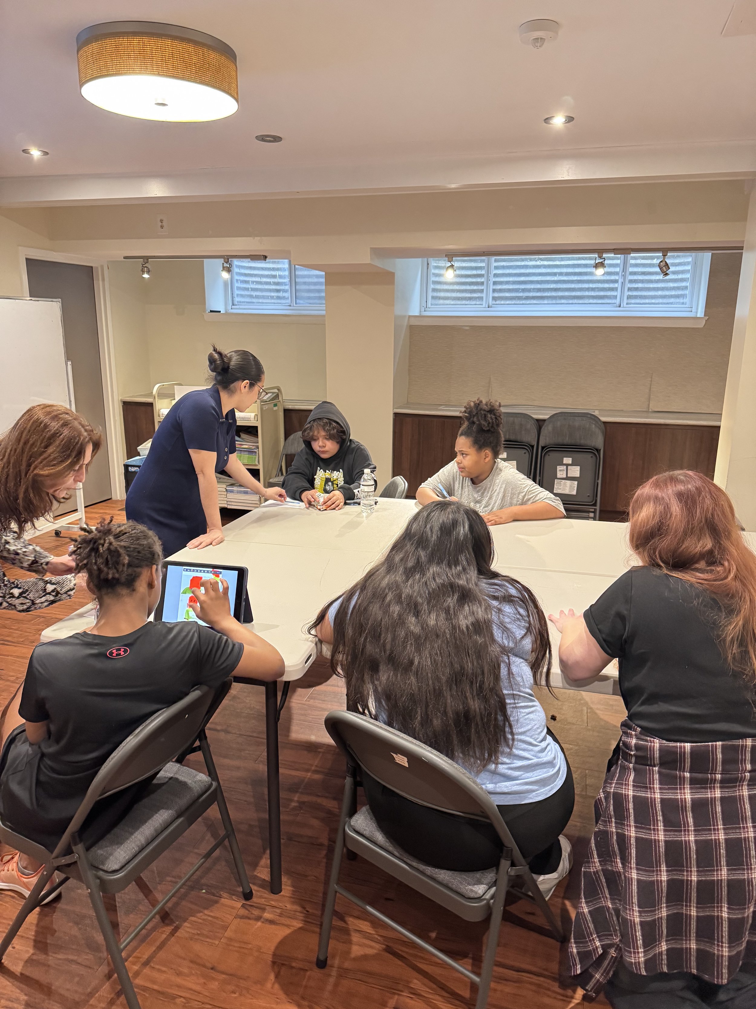 Group of children and adults gathered around a table in a classroom or community center, with a woman standing and interacting with a young boy in a hoodie. A girl in a black shirt is playing with a tablet, and others are seated and engaged.