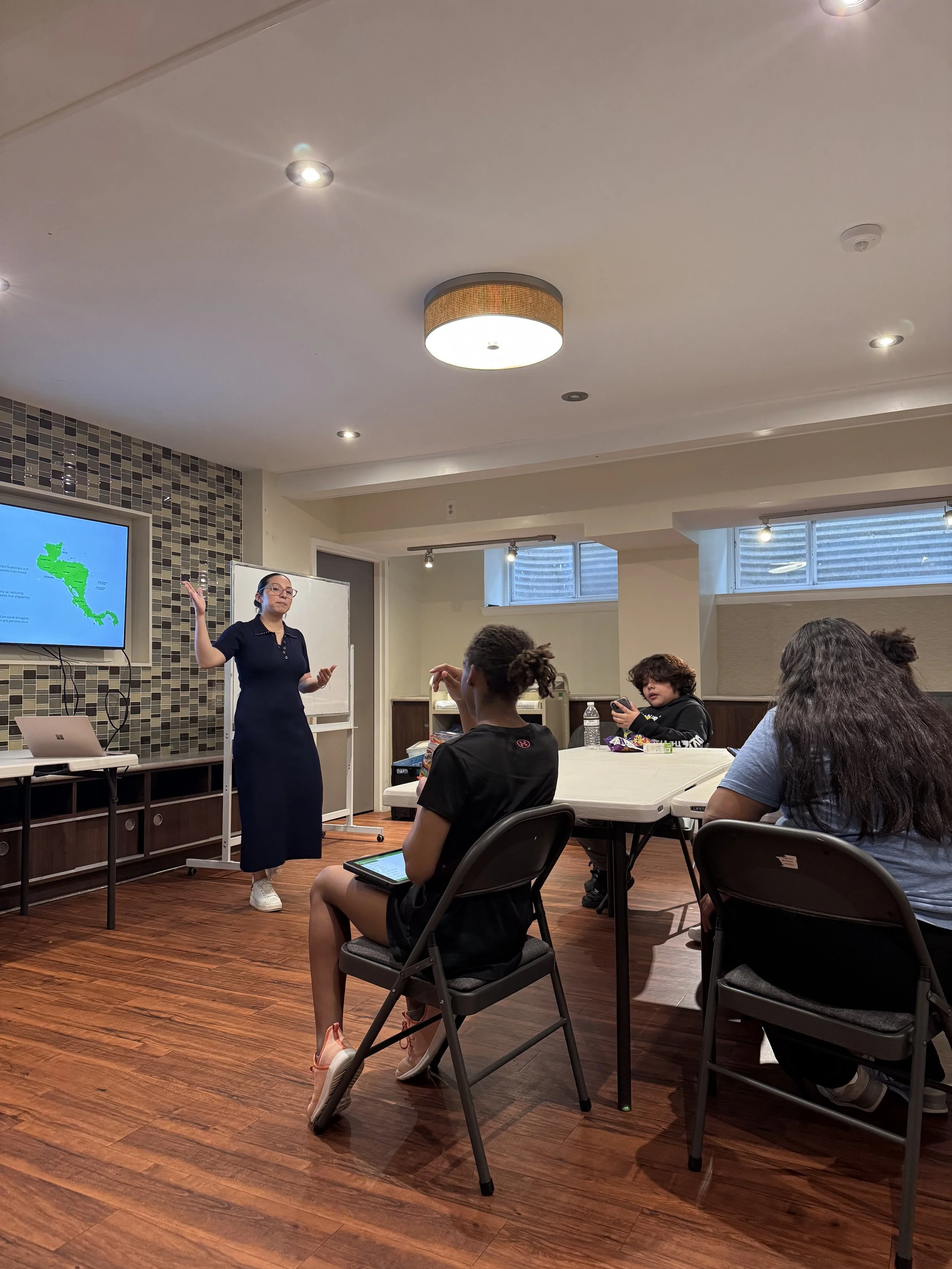 A woman in a black dress stands at the front of a room with several people seated, giving a presentation with a large screen displaying a map of Central America.
