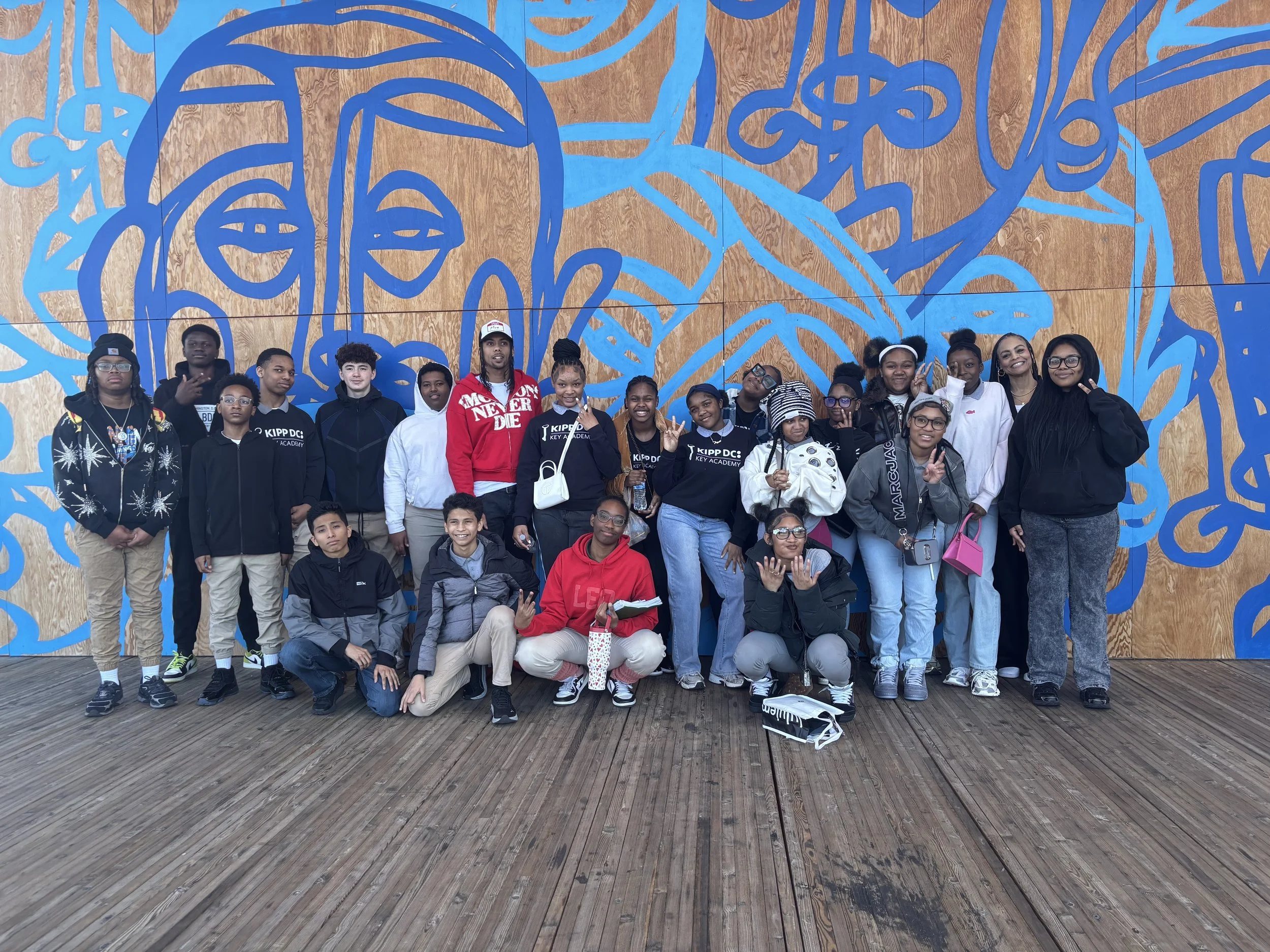 Group of diverse students standing and sitting in front of a large wooden panel wall with abstract blue artwork. Some students are smiling and making peace signs.