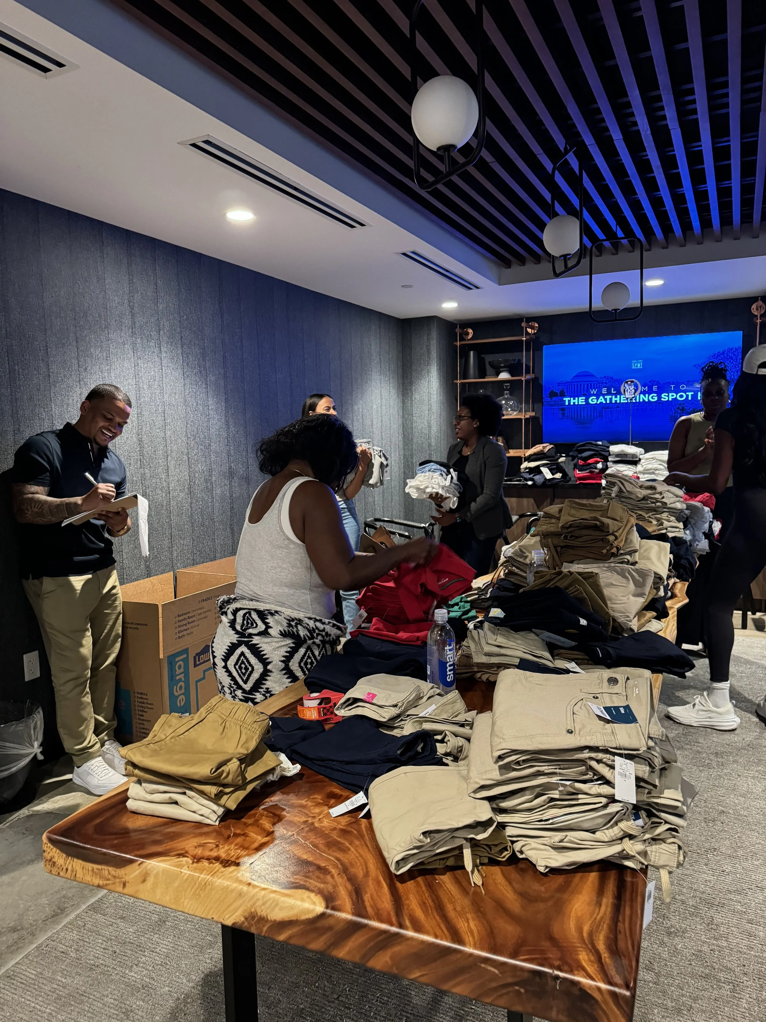 A group of people in a store or donation room sorting and organizing clothes on tables. The tables are filled with folded clothing items, including khaki pants, shirts, and other garments. There is a large flat-screen television on the wall displaying 'WELCOME TO THE GATHERING SPOT.' The room has a modern design with a textured gray wall, wooden shelving, and black ceiling slats with hanging light fixtures.