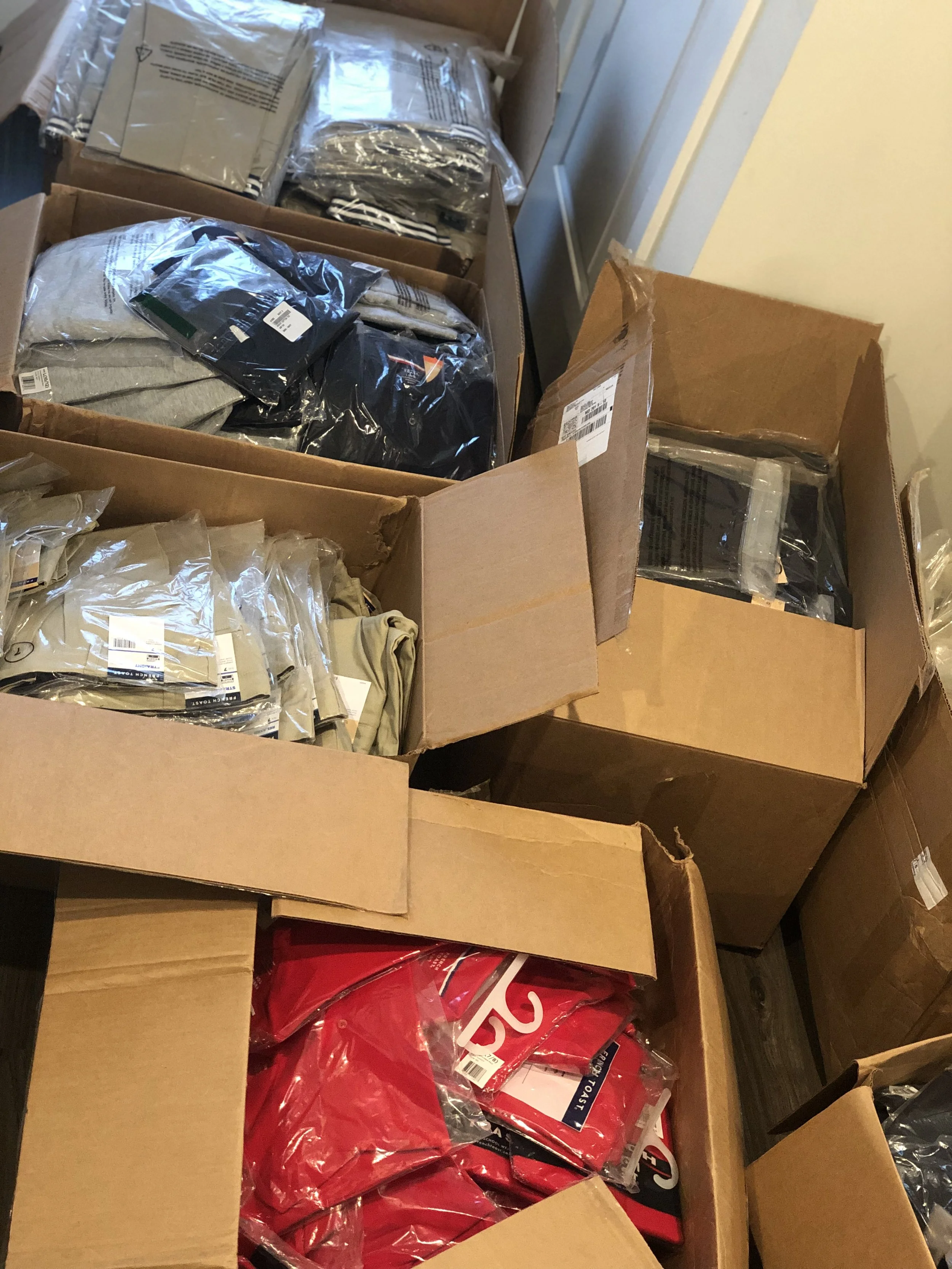 Open cardboard boxes filled with packaged items, including black, grey, and red plastic bags, with some items labeled, stacked inside a room with a door and yellow walls.