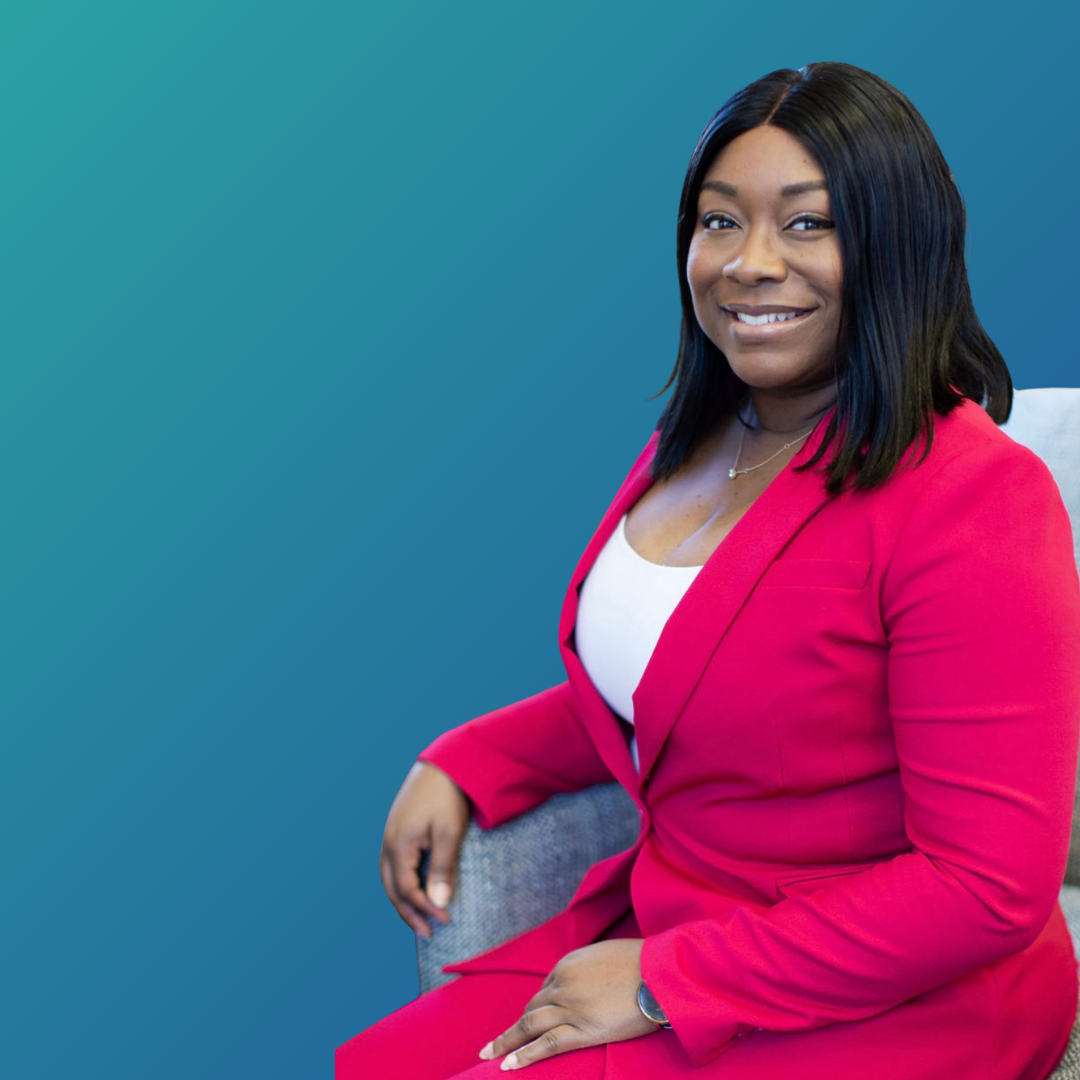 A woman with shoulder-length black hair wearing a red blazer and white top, sitting on a gray chair against a blue gradient background, smiling.