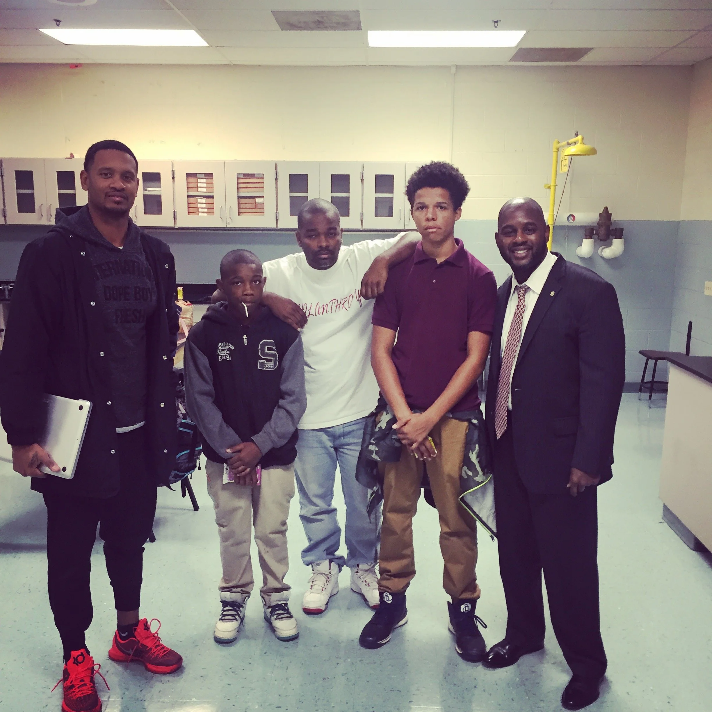 A group of five men and boys standing together in a school or community room. They are facing the camera, some with arms around each other. The background shows yellow and white cabinets, a yellow lamp, and plumbing pipes on the wall.