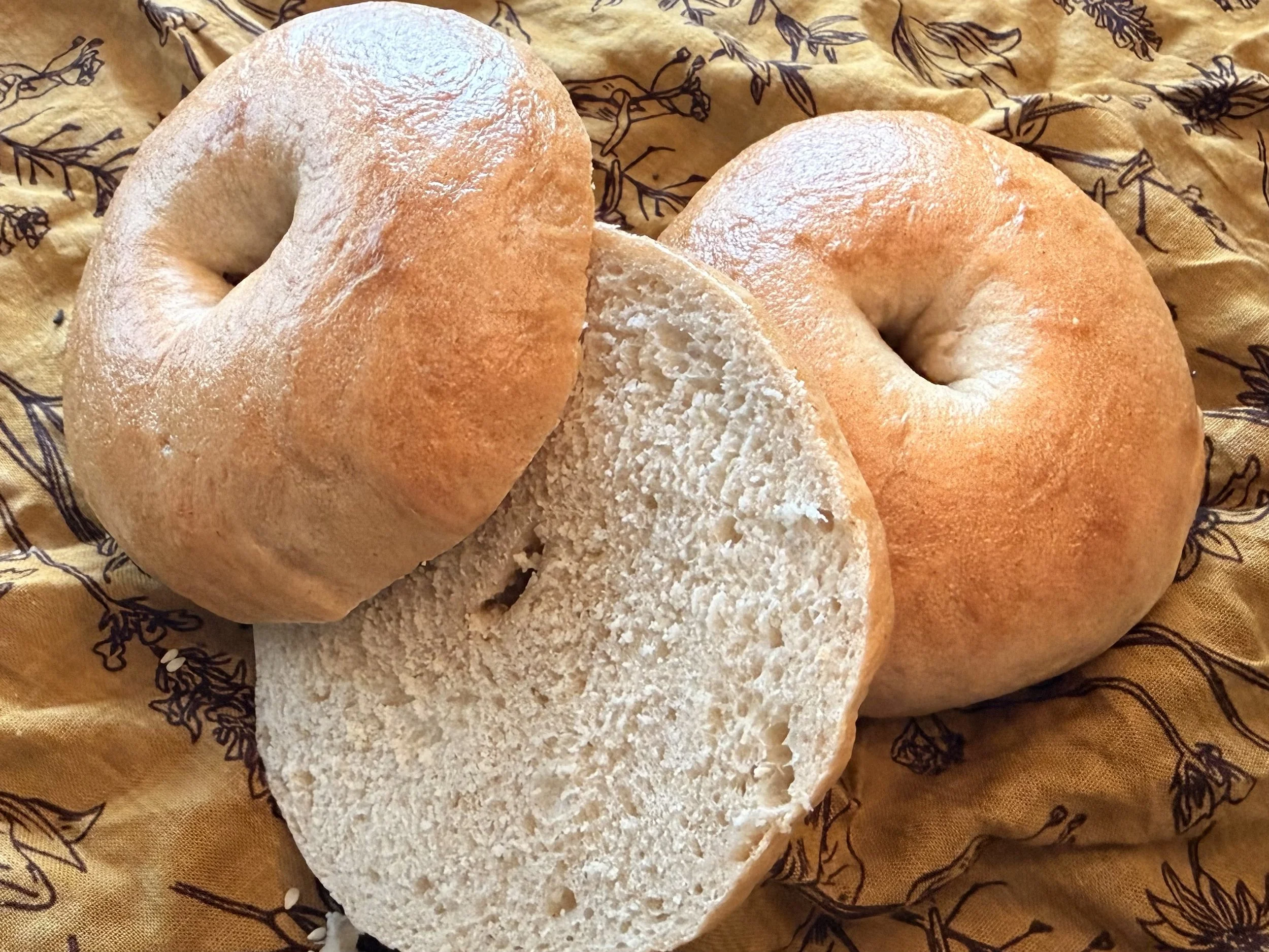 Two round, golden-brown baked bread rolls with a dusting of flour, one of which is partially sliced open, placed on a patterned cloth.