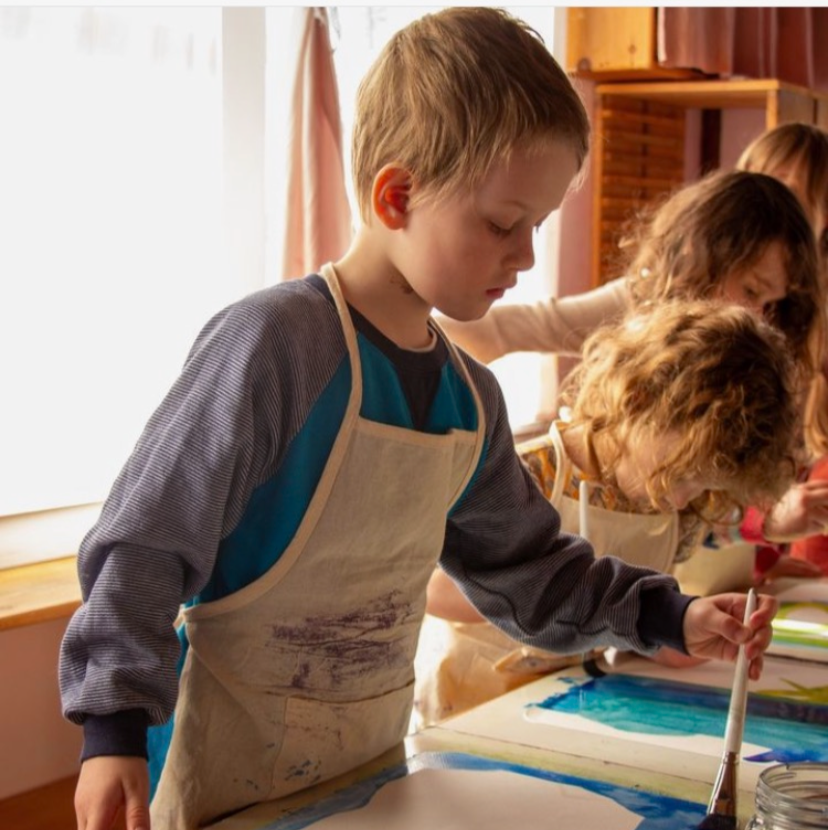 Children engaged in a watercolor painting activity on a table in a classroom setting, with sunlight coming through the window.