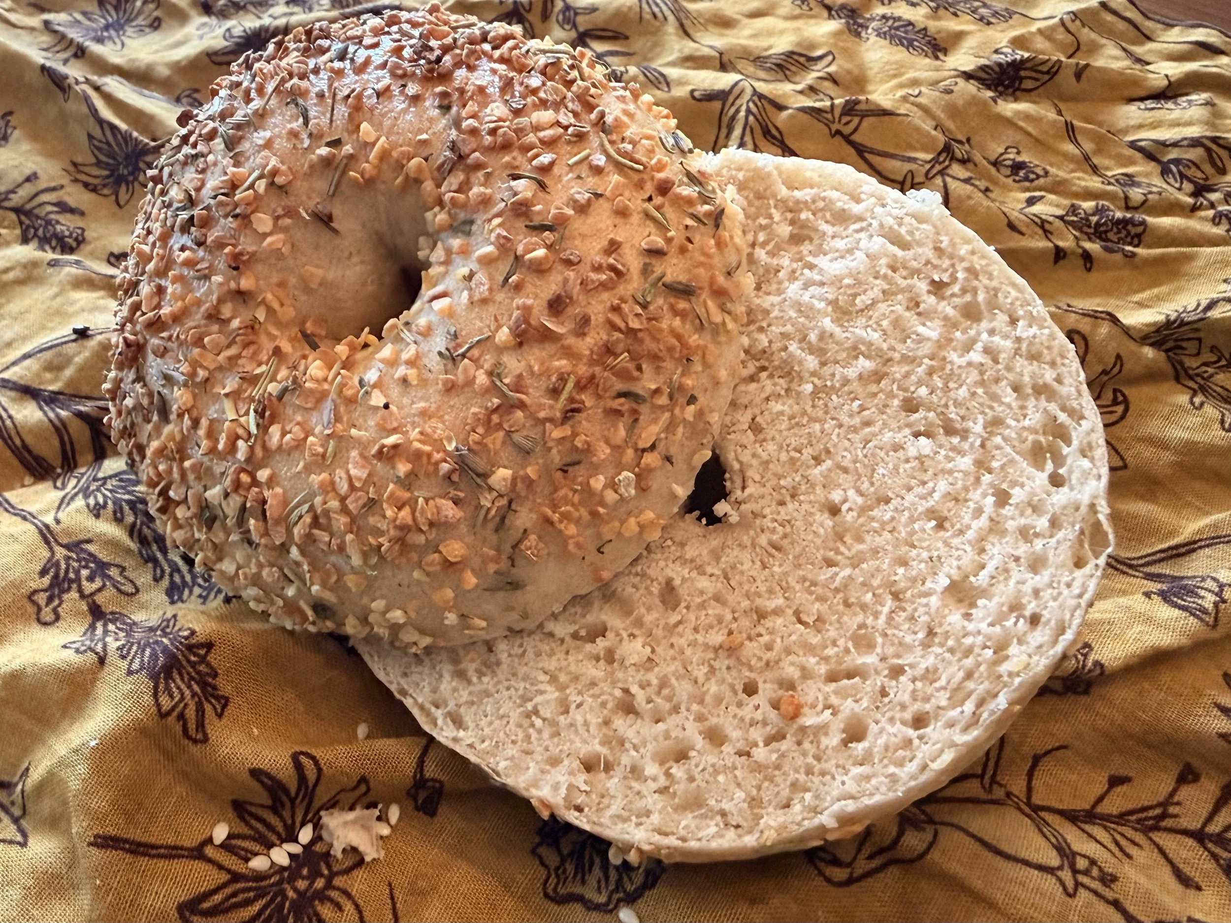 Round loaf of bread with a crust covered in chopped nuts and herbs, sliced to reveal a soft interior, on a patterned fabric surface.