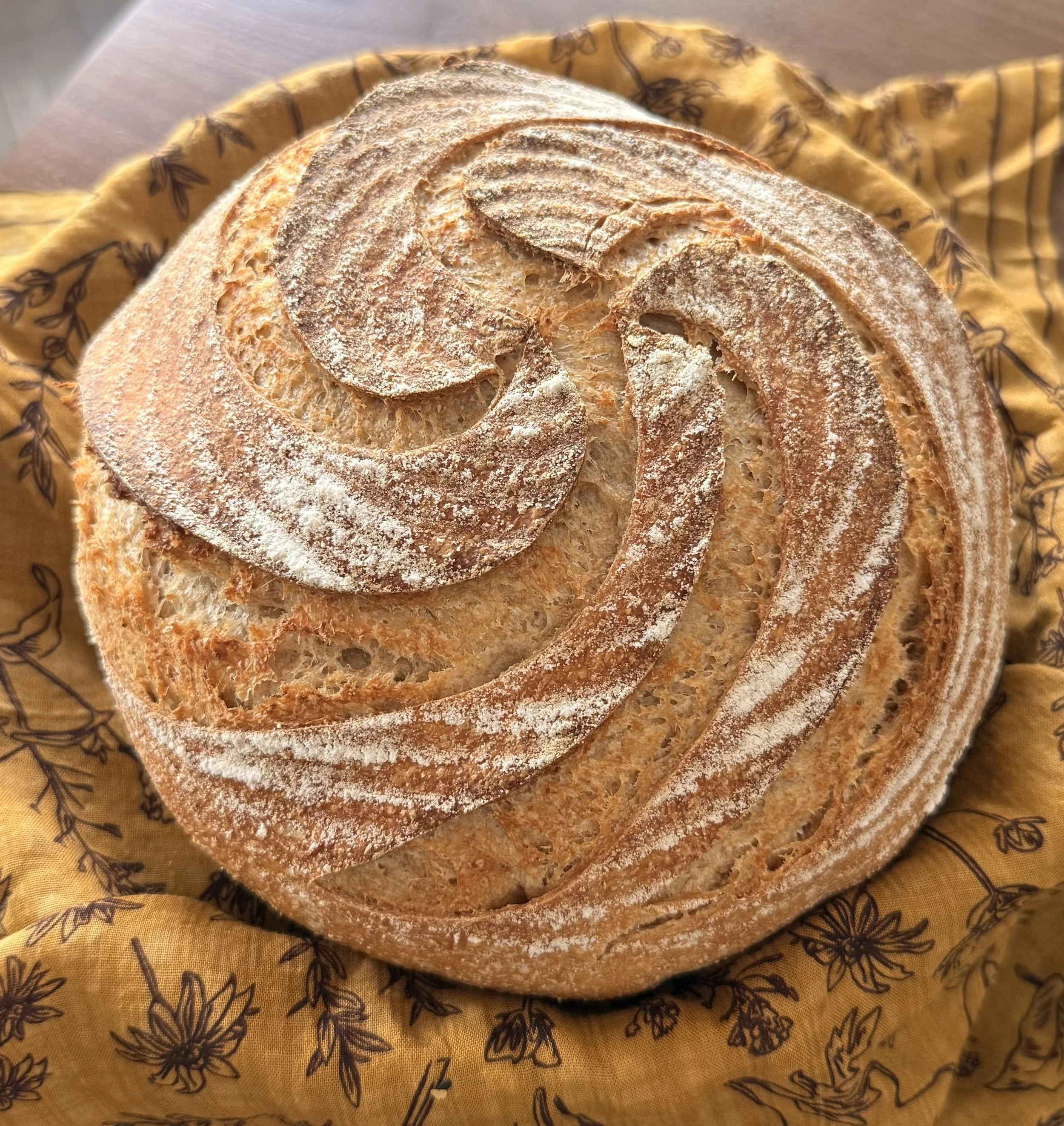 Close-up of sliced, round loaf of bread with a dusting of flour on top, resting on yellow cloth with black floral pattern.