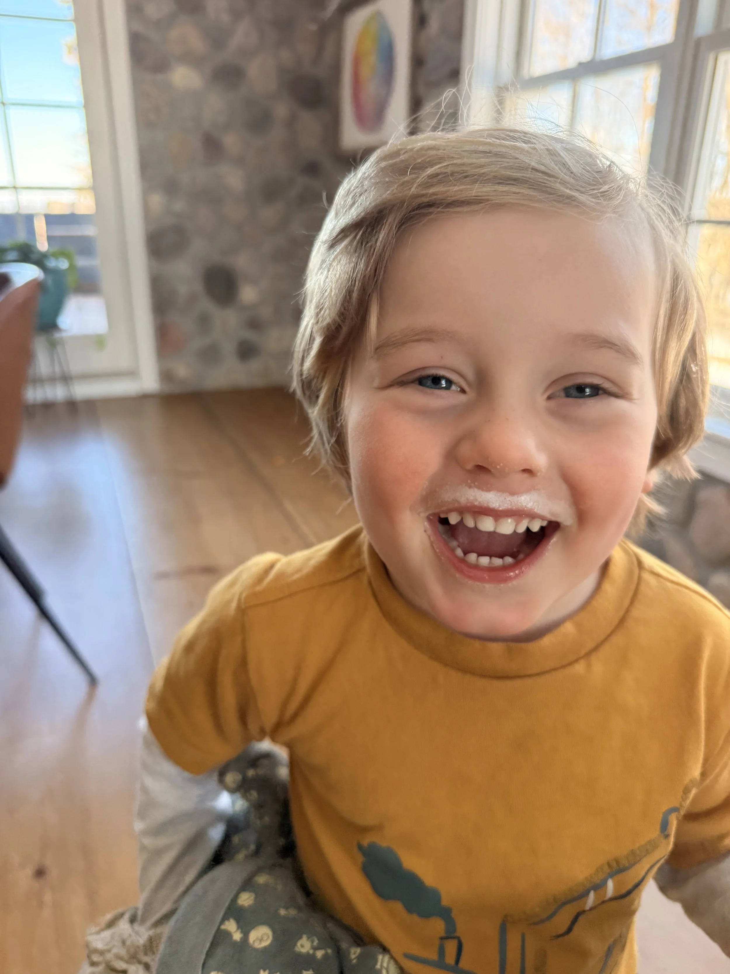 Close-up of a happy young boy with blonde hair and blue eyes, smiling with toothpaste on his face, wearing a mustard yellow shirt, indoors near a window with stone wall and colorful artwork in the background.