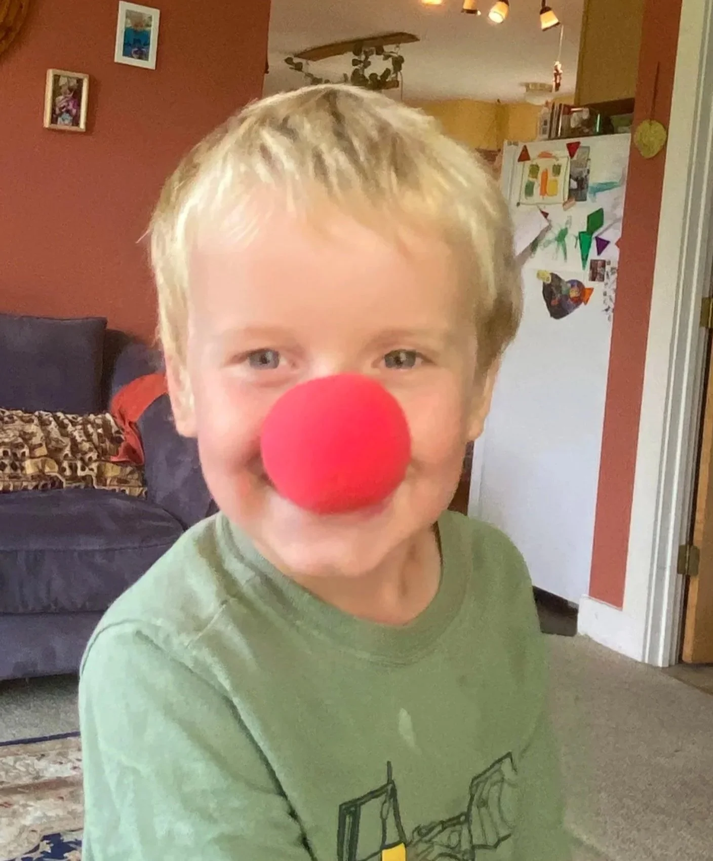 A young child with blond hair and blue eyes wearing a red clown nose and a green shirt, smiling in a cozy living room with artworks and a white refrigerator decorated with children's drawings.