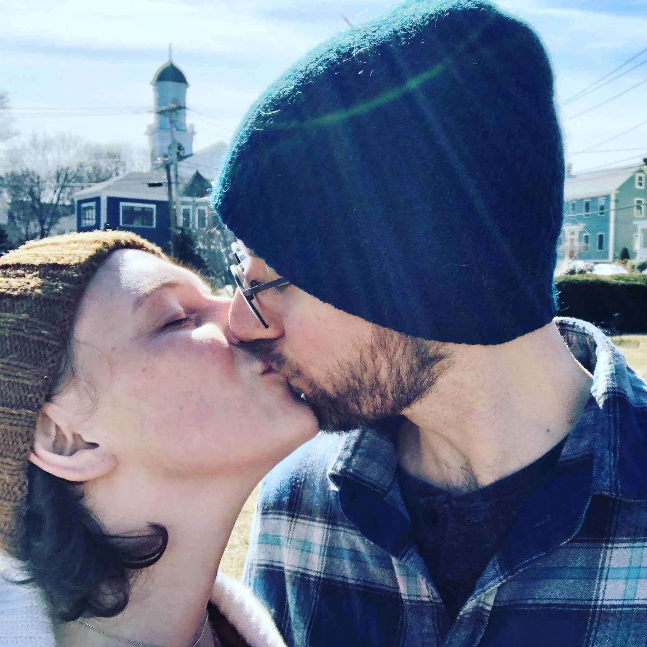 A woman and a man are close together, appearing to share a kiss. The woman has dark hair, and the man wears glasses and a dark beanie. They are outdoors with houses and a church tower visible in the background.