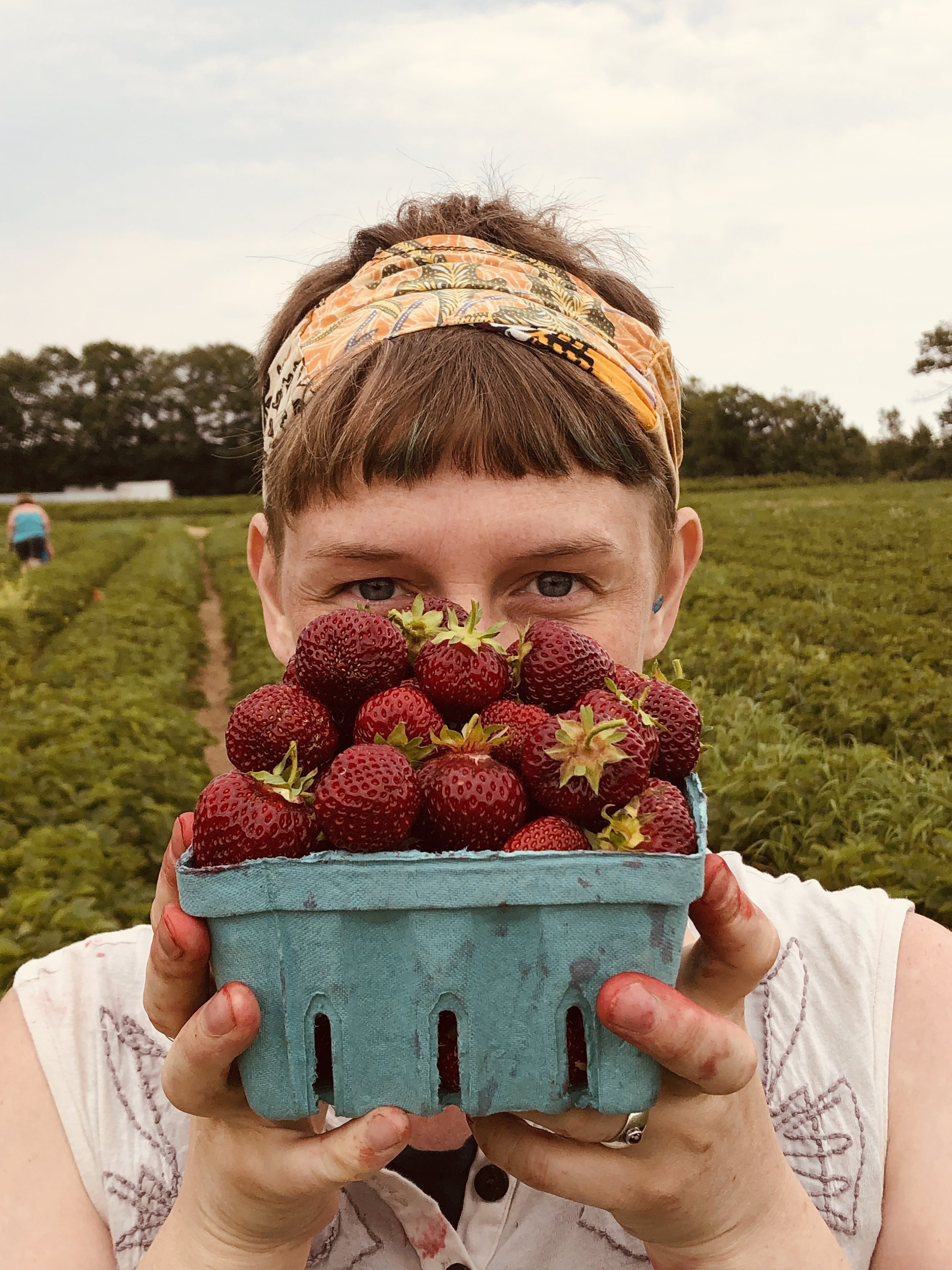 A person holding a basket of strawberries outdoors in a field, with another person visible in the background.