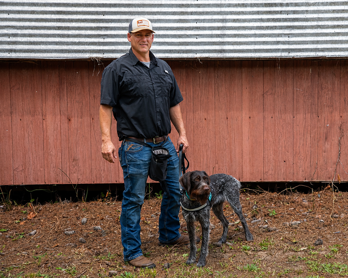 A man standing outdoors with a dog on a leash in front of a red wooden building, ground covered with pine needles and small pinecones.